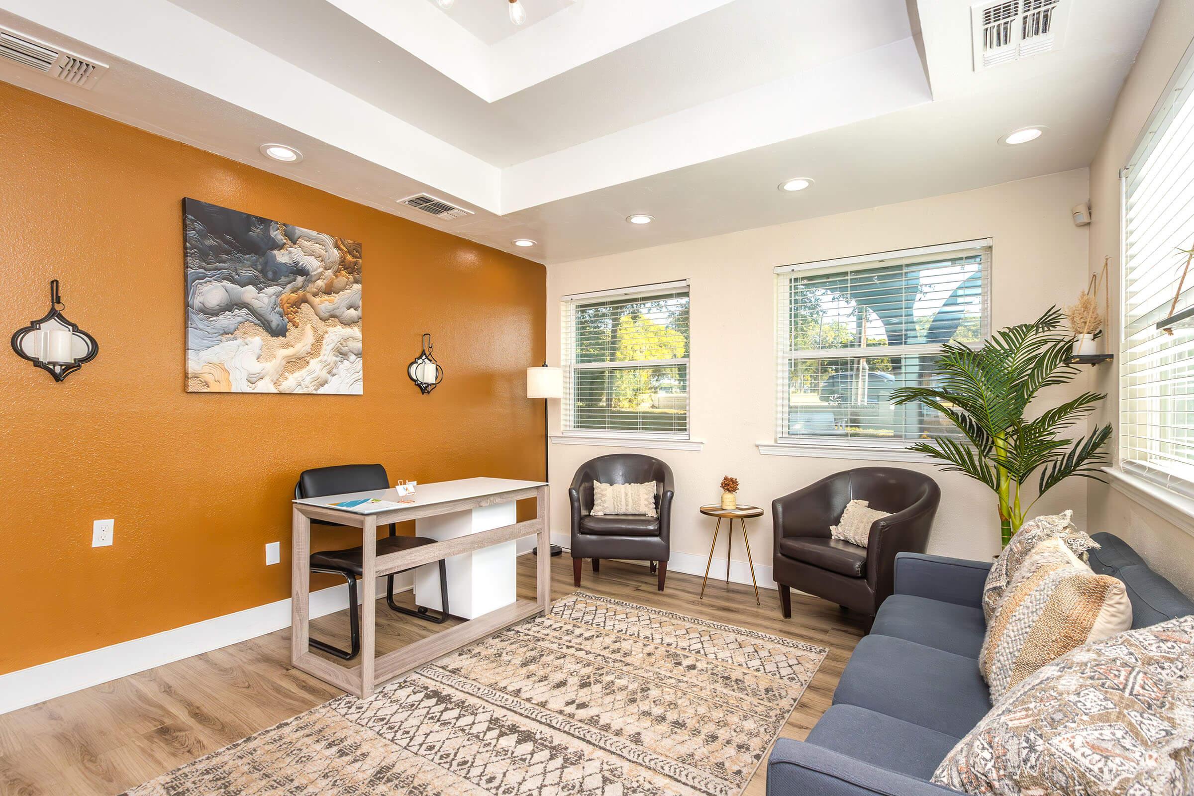 A modern, inviting office space featuring a brown accent wall, large windows, and plenty of natural light. The room includes a small desk with a chair, two comfortable armchairs, and a decorative rug. A piece of wall art adds color and interest, while indoor plants enhance the cozy atmosphere.