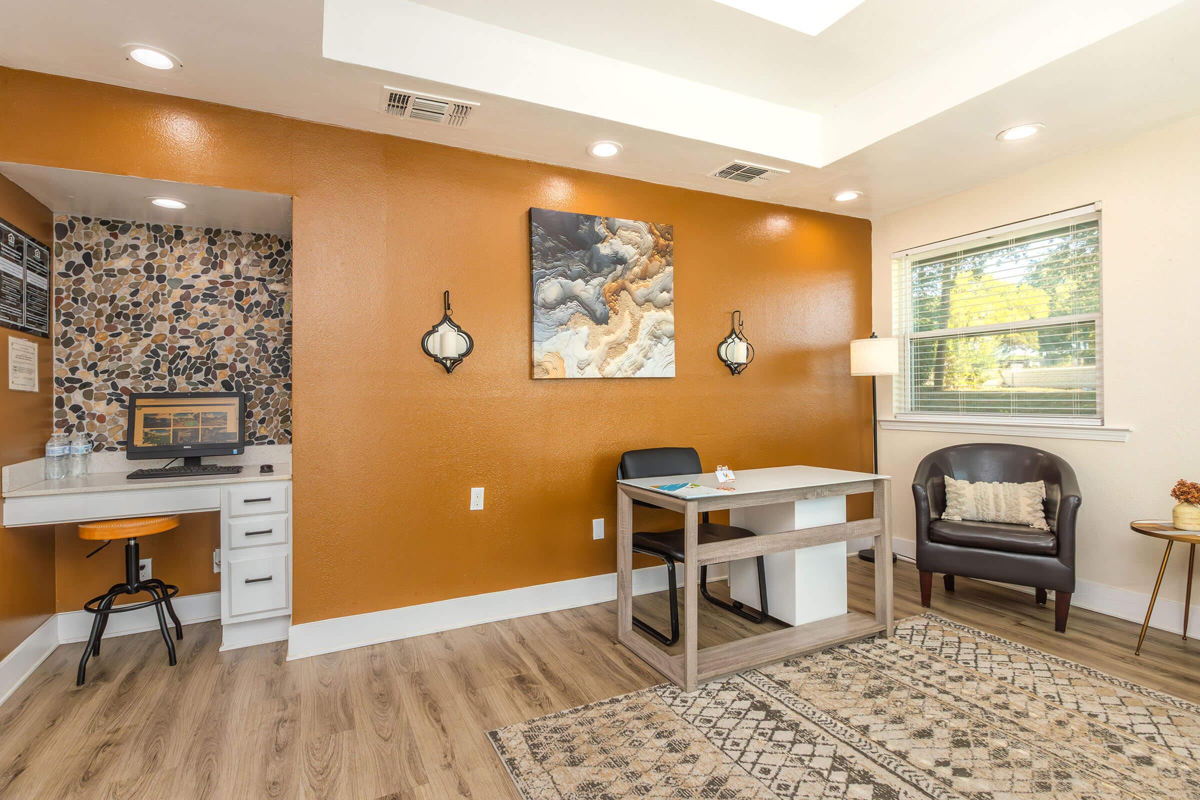A modern office space featuring an orange accent wall, a wooden desk, and a chair. There’s a cozy armchair in the corner, a patterned rug on the floor, and a small workspace with a computer. Natural light comes through a window, enhancing the inviting atmosphere. Wall art adds a decorative touch.