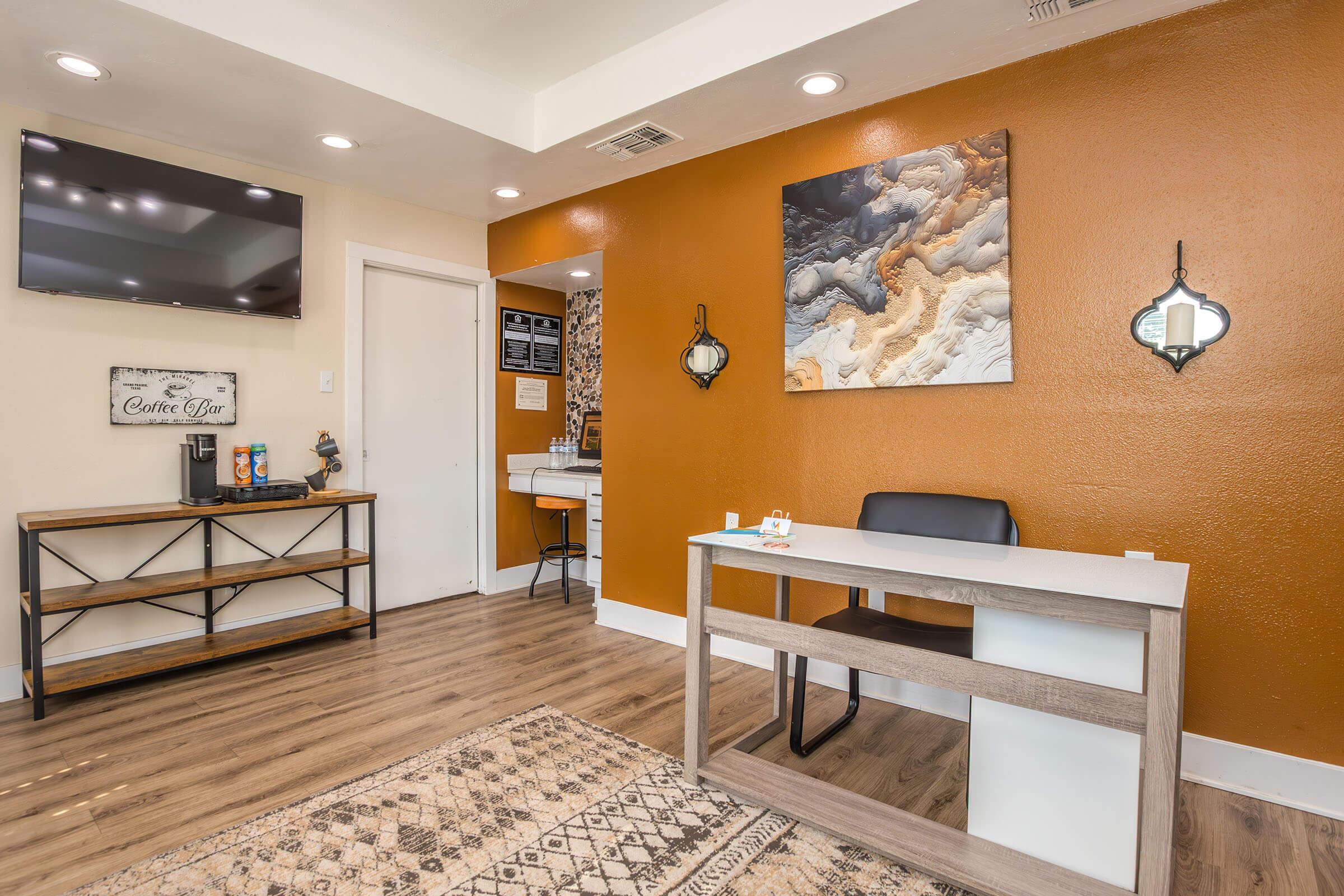 A modern, cozy reception area featuring a desk and a chair, with a brown accent wall and decorative wall art. A television is mounted on the wall, and a patterned rug adds warmth. A small coffee area is visible, along with additional decorations and exam rooms in the background.