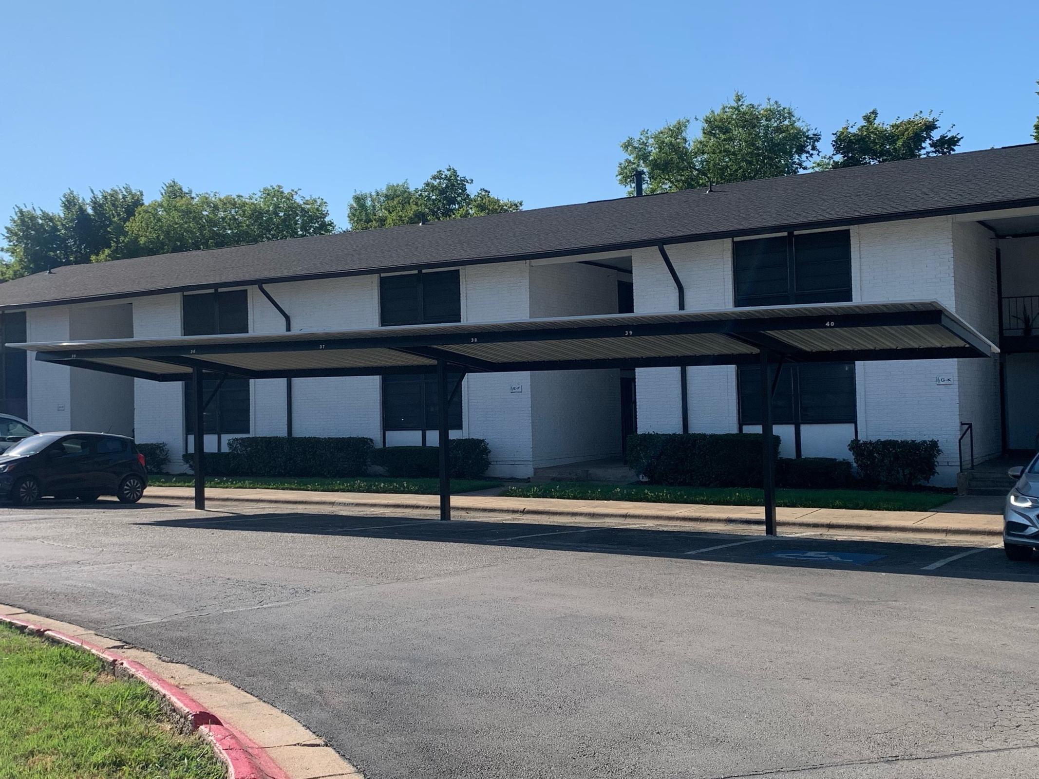 A view of a parking area with a covered carport in front of a residential building. The building features a light exterior with multiple windows, and the parking area is partially shaded by the carport structure. Clear blue sky and trees are visible in the background.