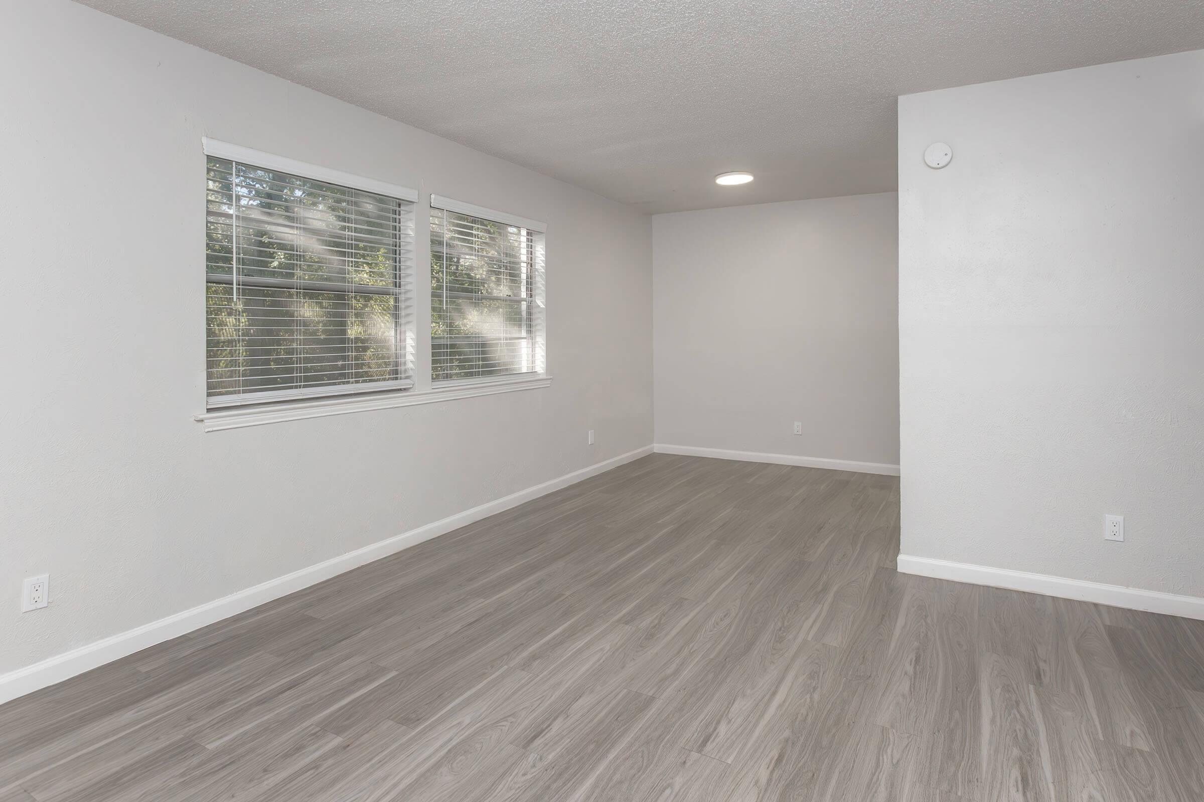 A spacious, empty room with light gray walls and a smooth, light wood-like floor. There are two large windows with blinds allowing natural light to enter. The ceiling features a central light fixture, and one corner has a small, round wall-mounted object. The overall atmosphere is bright and inviting.