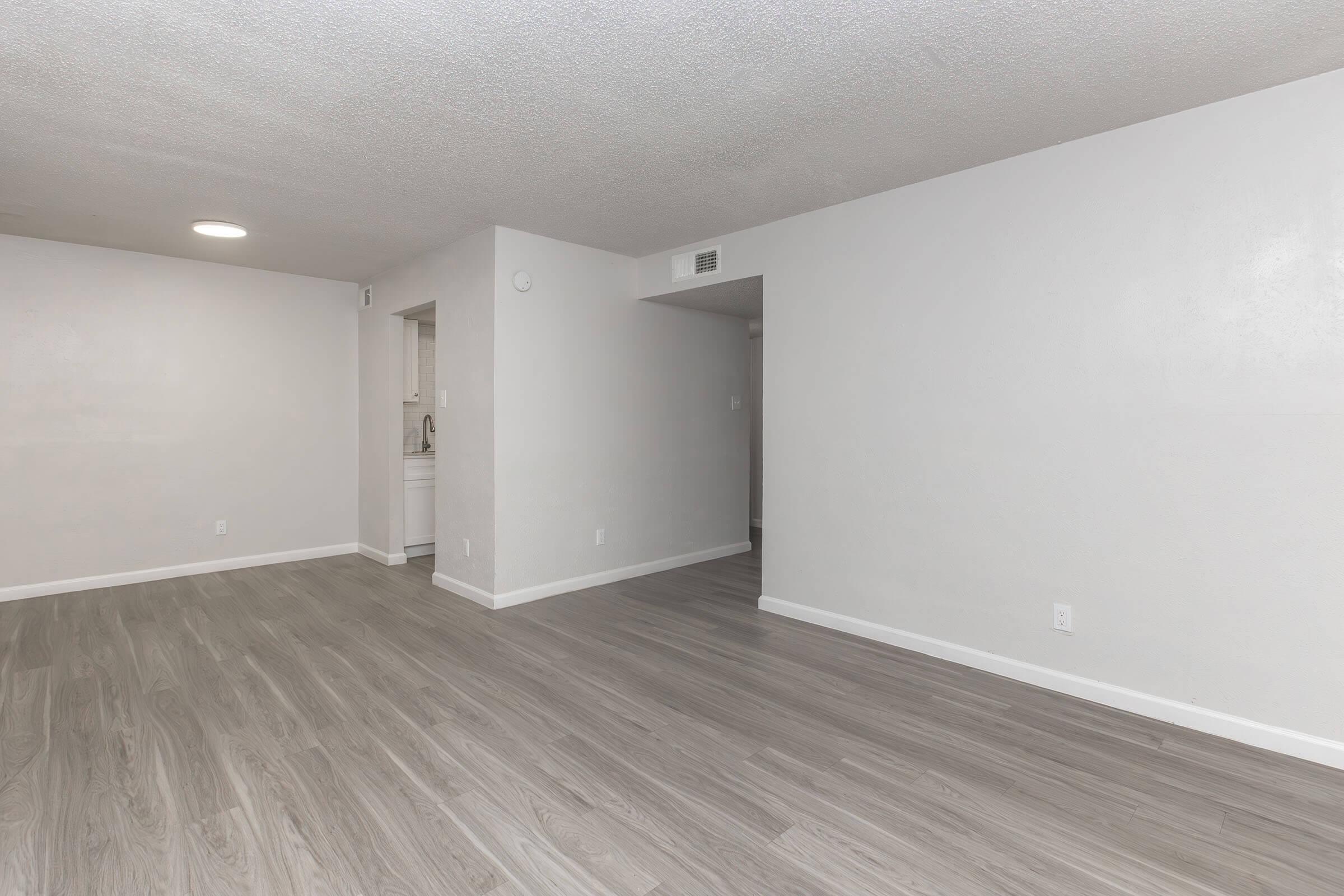 A spacious, empty room with light gray walls and a smooth, light wood-like floor. To the left, a doorway leads to a kitchen area, while the walls are clean and unadorned, enhancing the airy feel of the space.