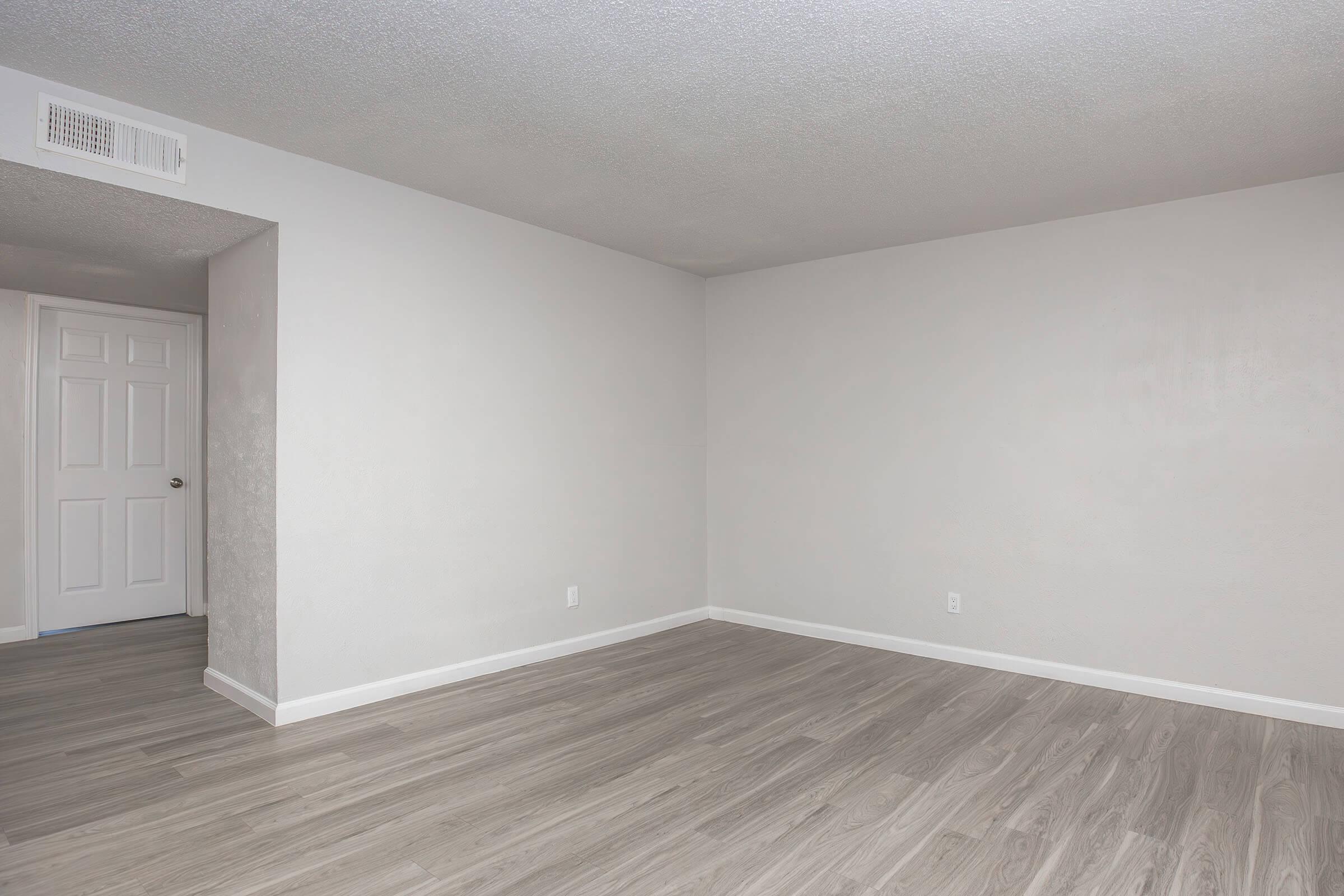 A spacious, empty room with light gray walls and wooden flooring. There is a doorway visible on the left leading to another room, and the ceiling has a textured finish. The overall ambiance is bright and neutral, making it suitable for various decorating styles.