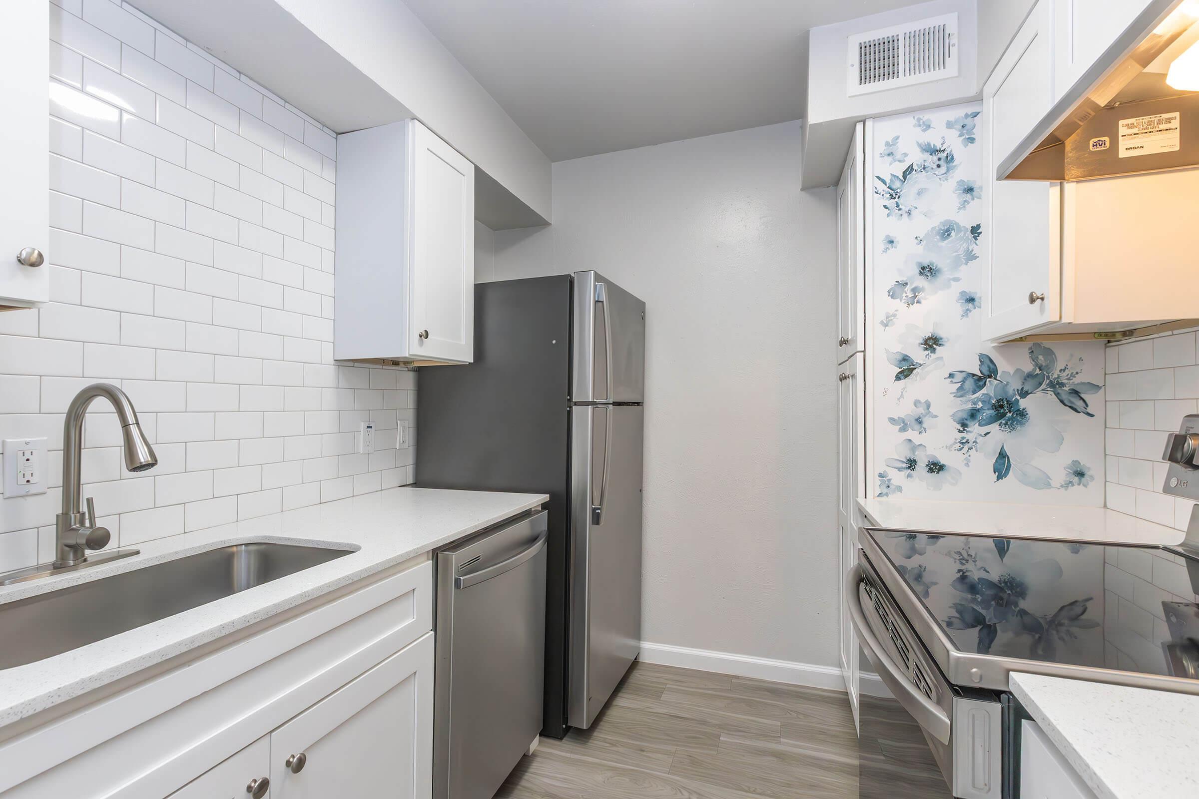 Modern kitchen featuring white cabinetry, a stainless steel refrigerator, a sink, and an oven with a stove. The backsplash is adorned with white tiles and a floral design in blue, while the walls are painted a light gray. Natural light enhances the clean and contemporary look of the space.