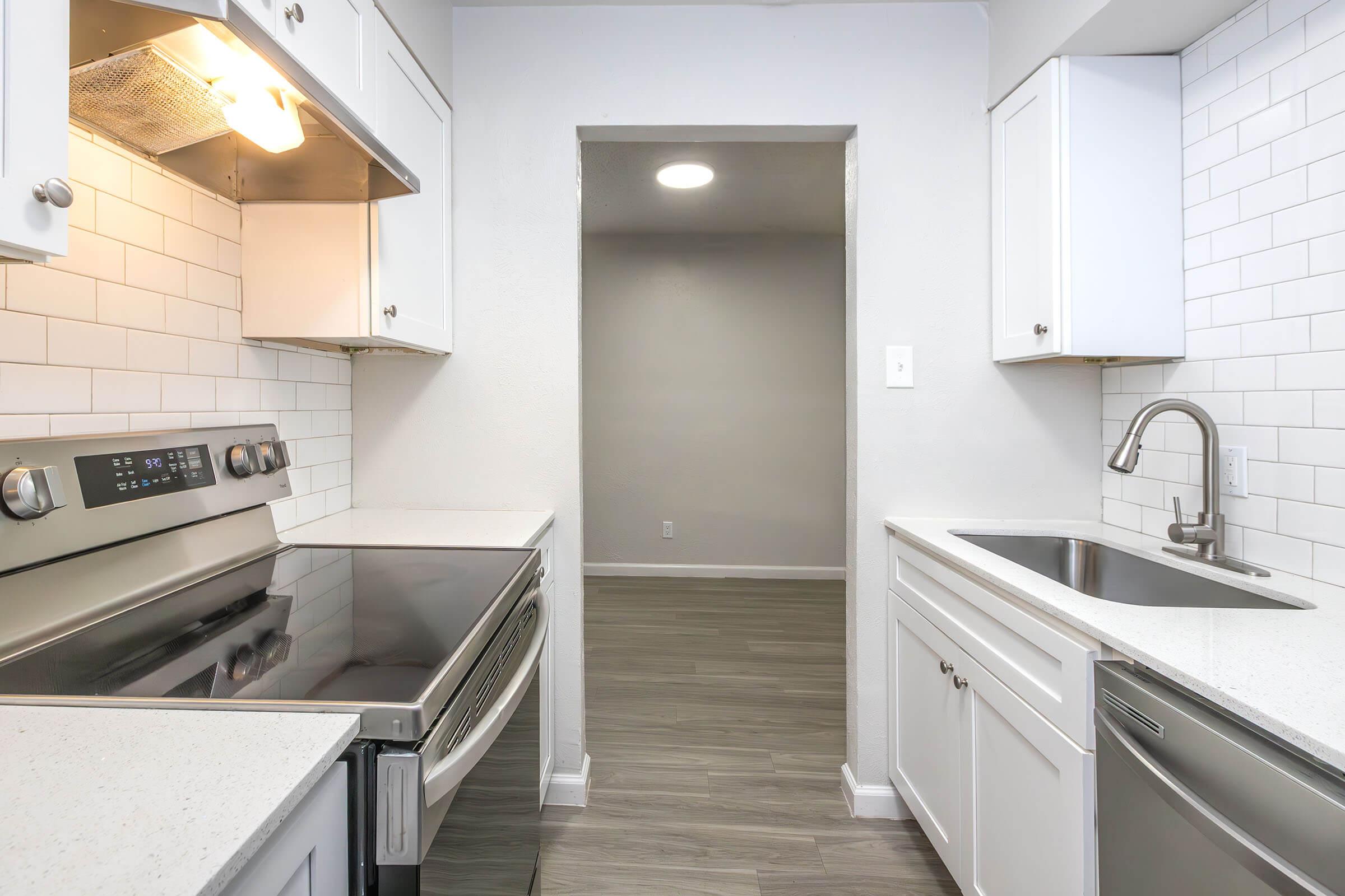 Modern kitchen with white cabinets, stainless steel appliances, and a subway tile backsplash. Features an electric range and a sink with a modern faucet. The room has gray wood-style flooring and leads to a doorway with a neutral-colored wall. Natural light from the ceiling fixture enhances the space.