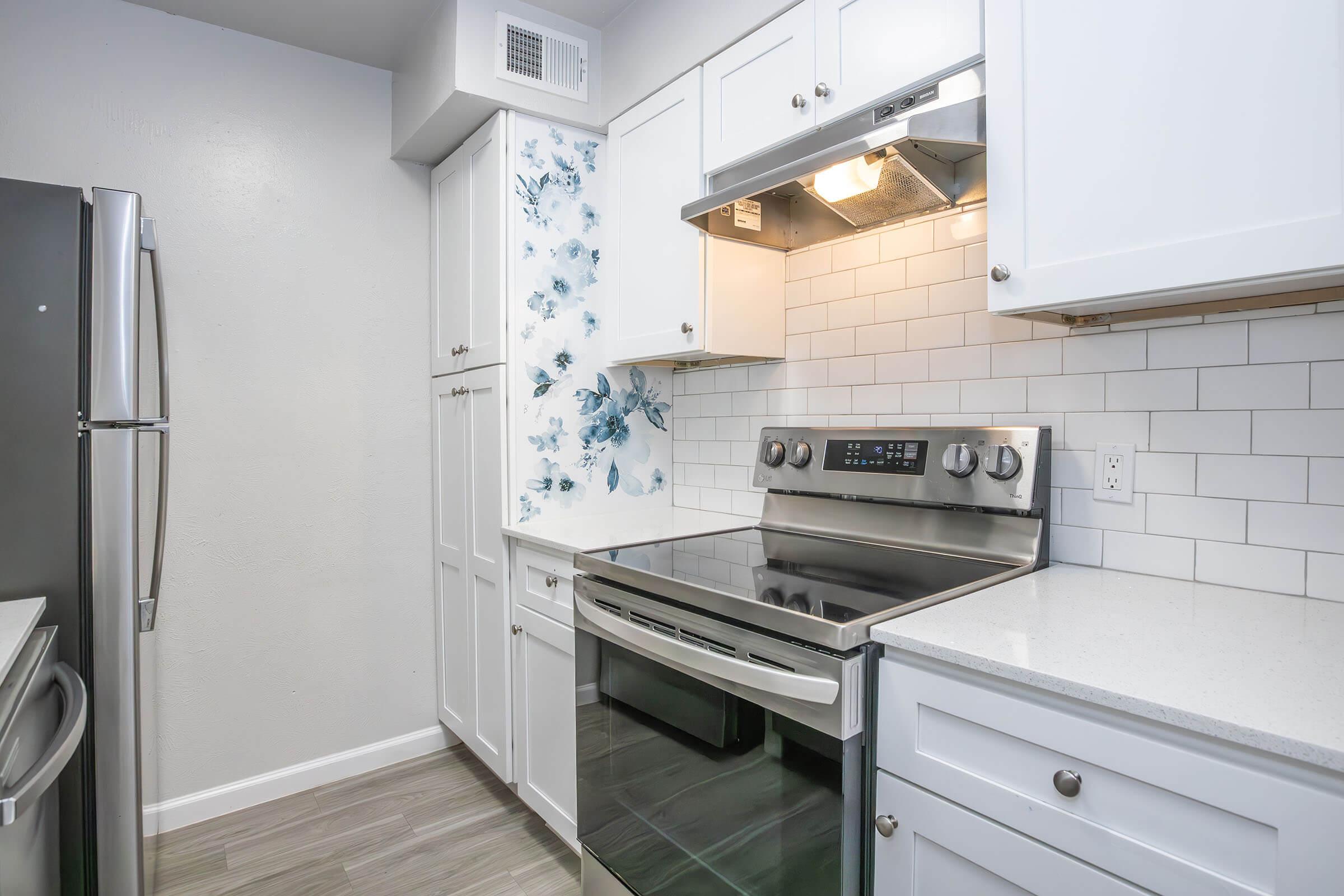 Modern kitchen featuring stainless steel appliances, including an oven and refrigerator, with white cabinetry and a blue accent wall. The space is bright and airy, with a combination of tile and hardwood flooring. Natural light enters through a window above the sink area.