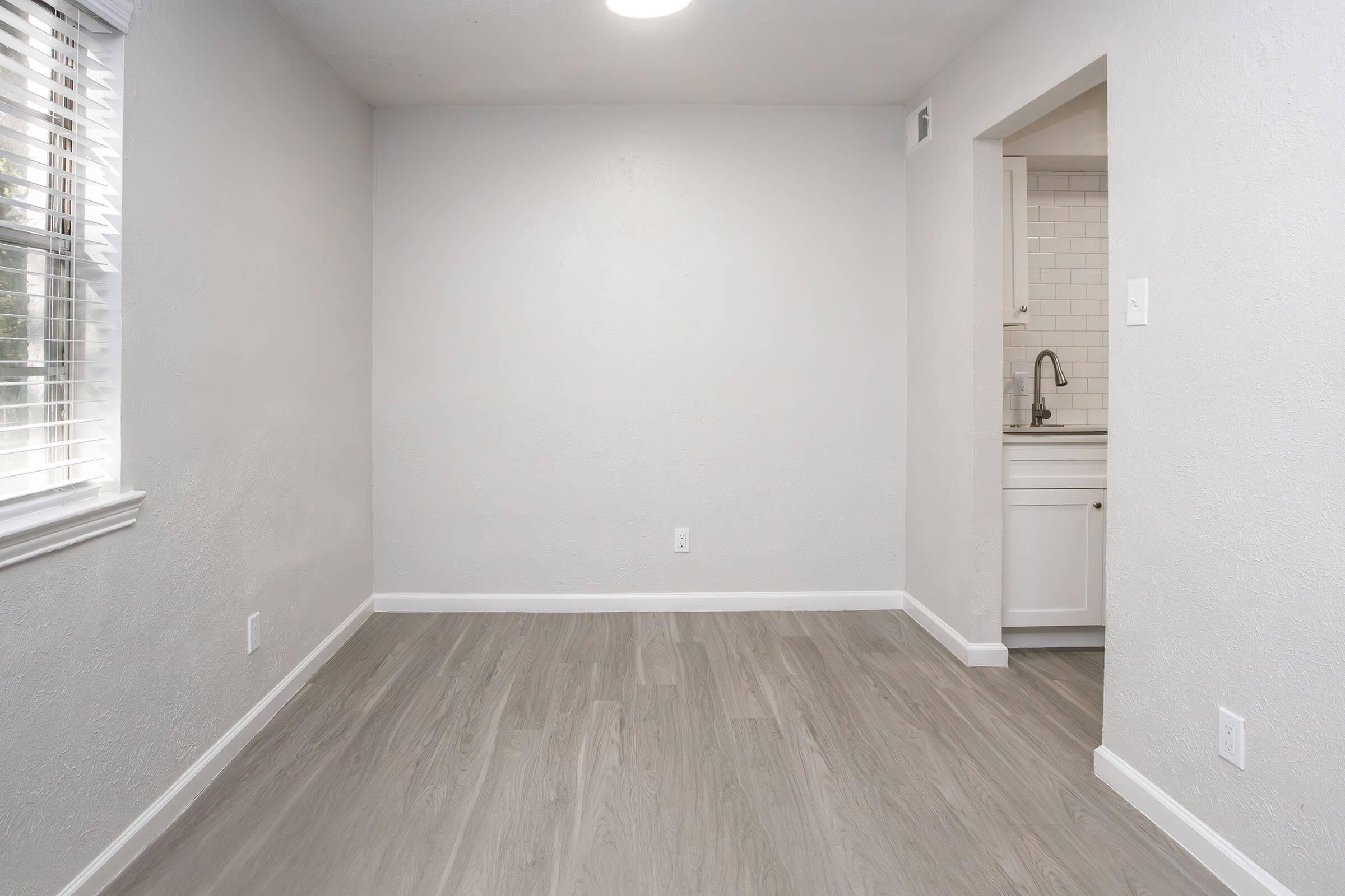 A small, empty room with light gray walls and a light-colored laminate floor. There is a window with white blinds on one side and an open doorway leading to a small kitchen area with white cabinets and a sink. The room is well-lit with natural light.