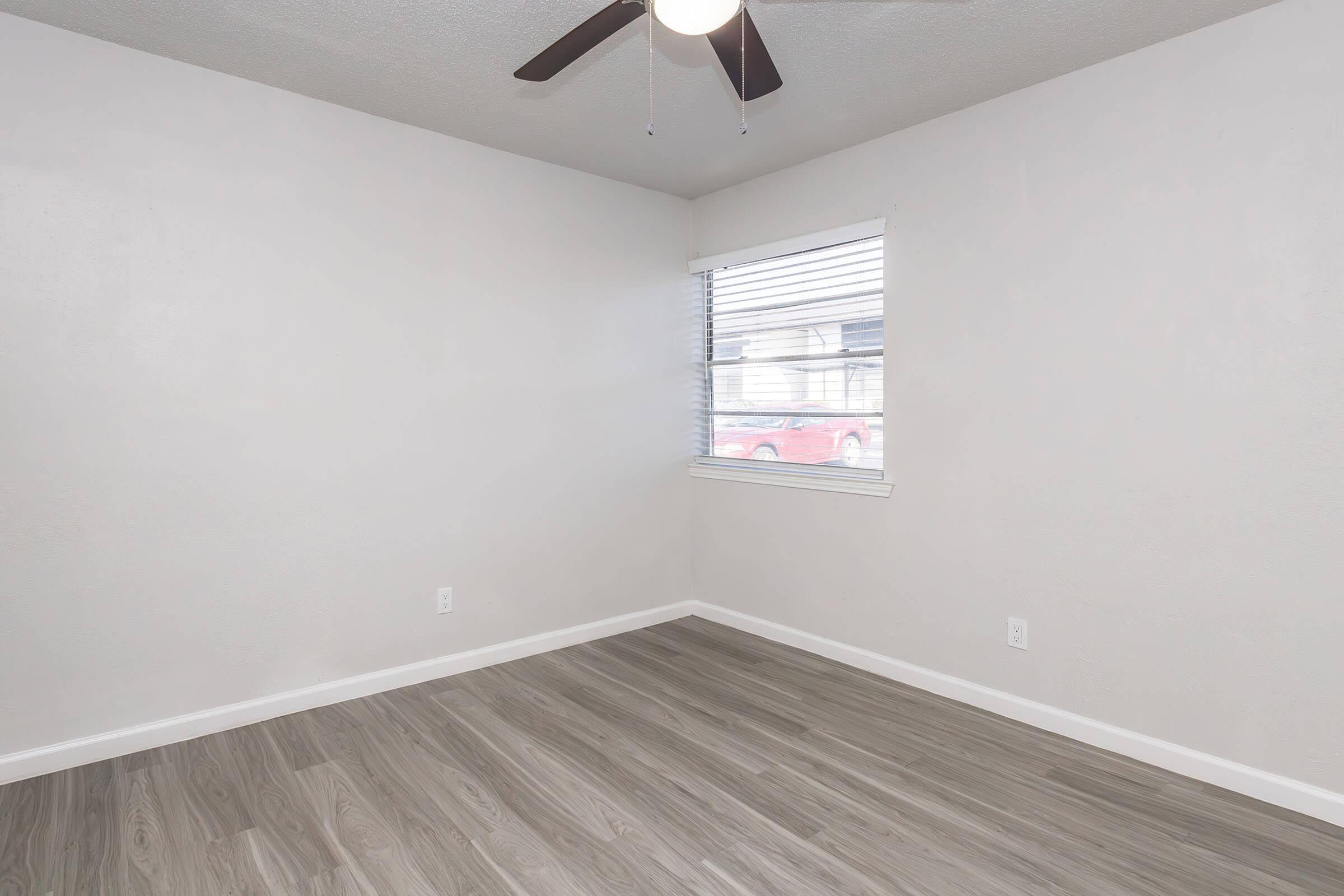 A vacant room with light gray walls, a ceiling fan, and a window with blinds. The floor is covered in light-colored wood laminate. Natural light enters through the window, offering a view of a car parked outside. The space is empty, creating a minimalist aesthetic.