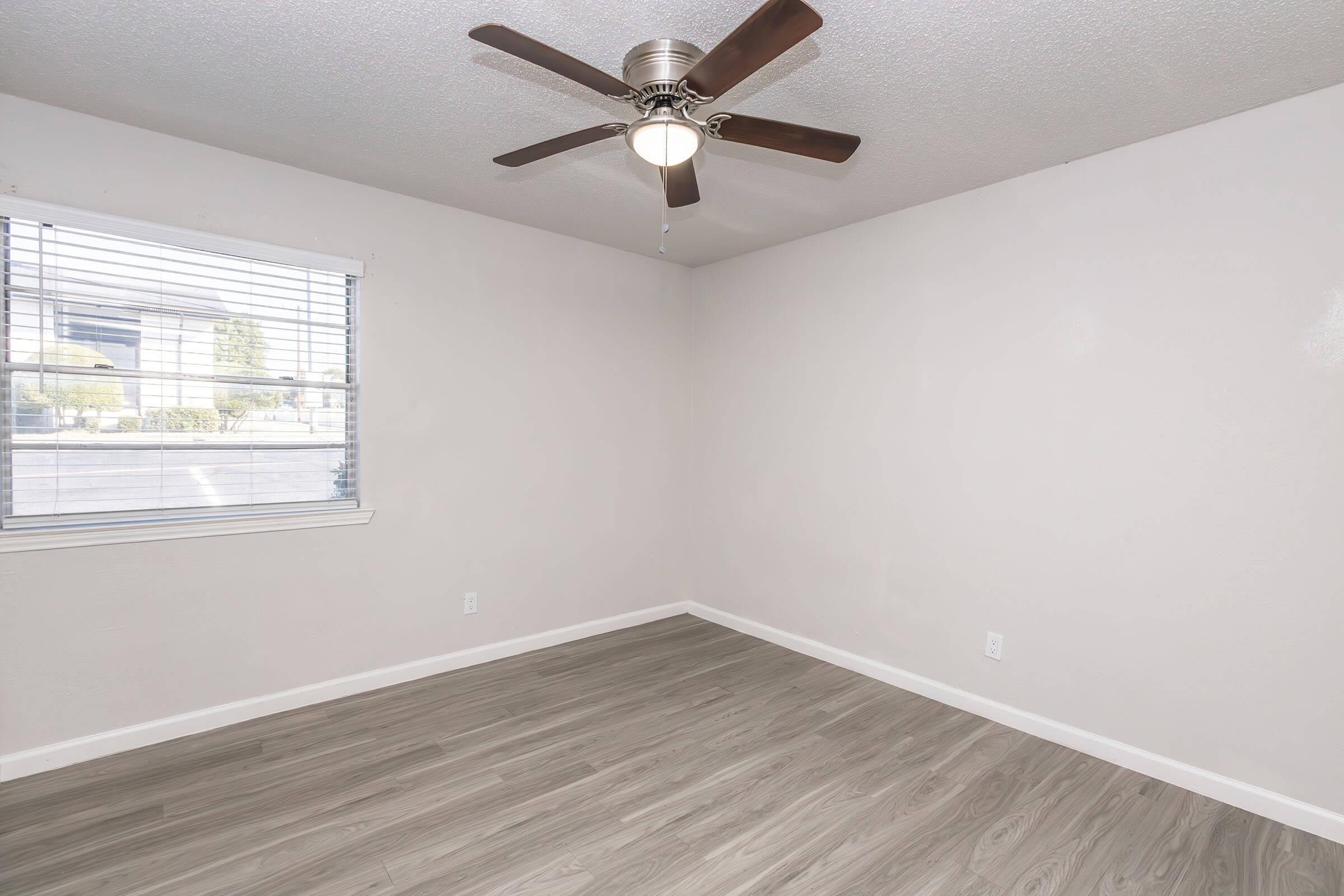 Empty room with light-colored walls and a ceiling fan. There is a window on one side with blinds, allowing natural light to enter. The floor has a light wood finish, creating a clean and open atmosphere.