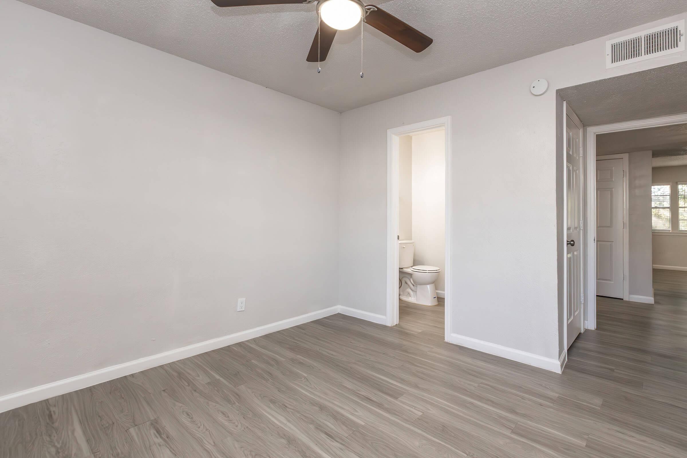 A spacious, empty room featuring light gray walls and a ceiling fan. The floor is covered with light brown laminate planks. A doorway leads to a bathroom on the left, while a second doorway is visible in the background. Natural light streams in from a window, illuminating the minimalist design and open layout.