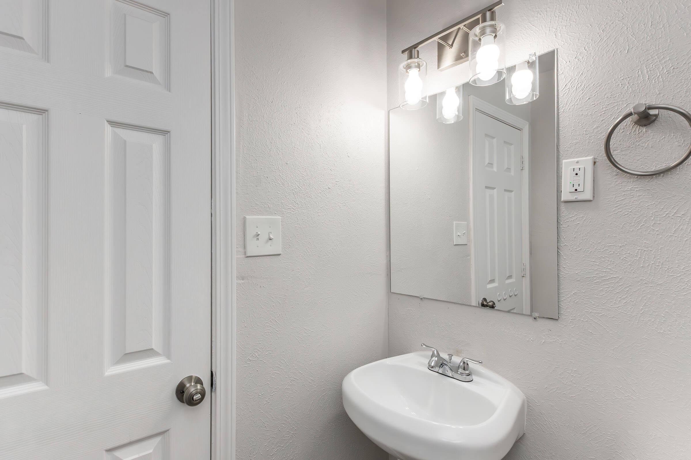 A clean bathroom with light gray walls. There is a white sink mounted on the wall, a large mirror above it, and a modern light fixture with three bulbs. A white door, partially closed, is visible in the background. The space is well-lit and minimalistic, creating a fresh and tidy appearance.