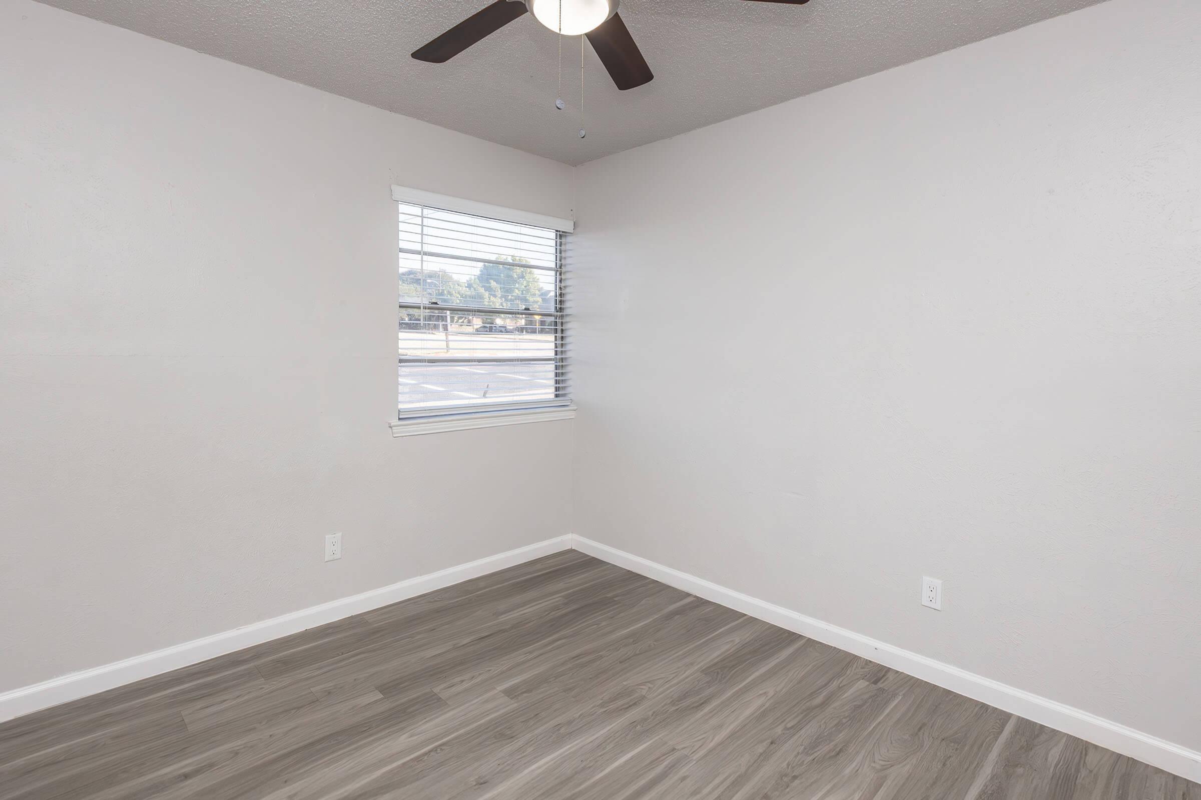 A small, empty room with light gray walls and a light wood-like floor. A ceiling fan is mounted on the ceiling, and there is a window with blinds allowing natural light to enter. The room appears clean and unfurnished, providing a blank space for potential use.