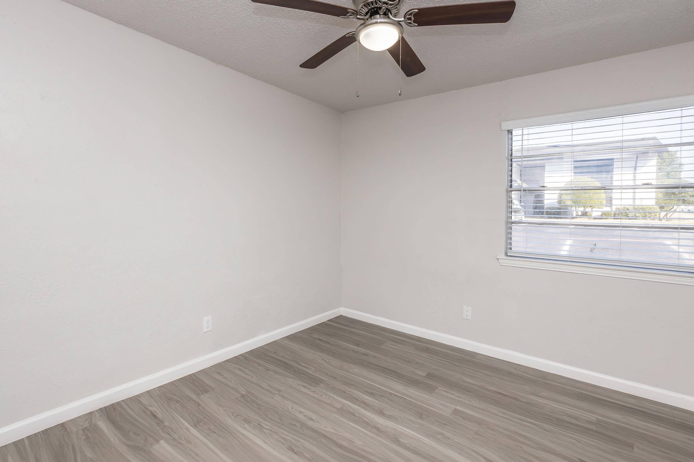Empty room with light-colored walls and a ceiling fan. There is a window with blinds on the right, allowing natural light to enter, and a light wood floor. The room is unfurnished and spacious, creating a minimalist atmosphere.