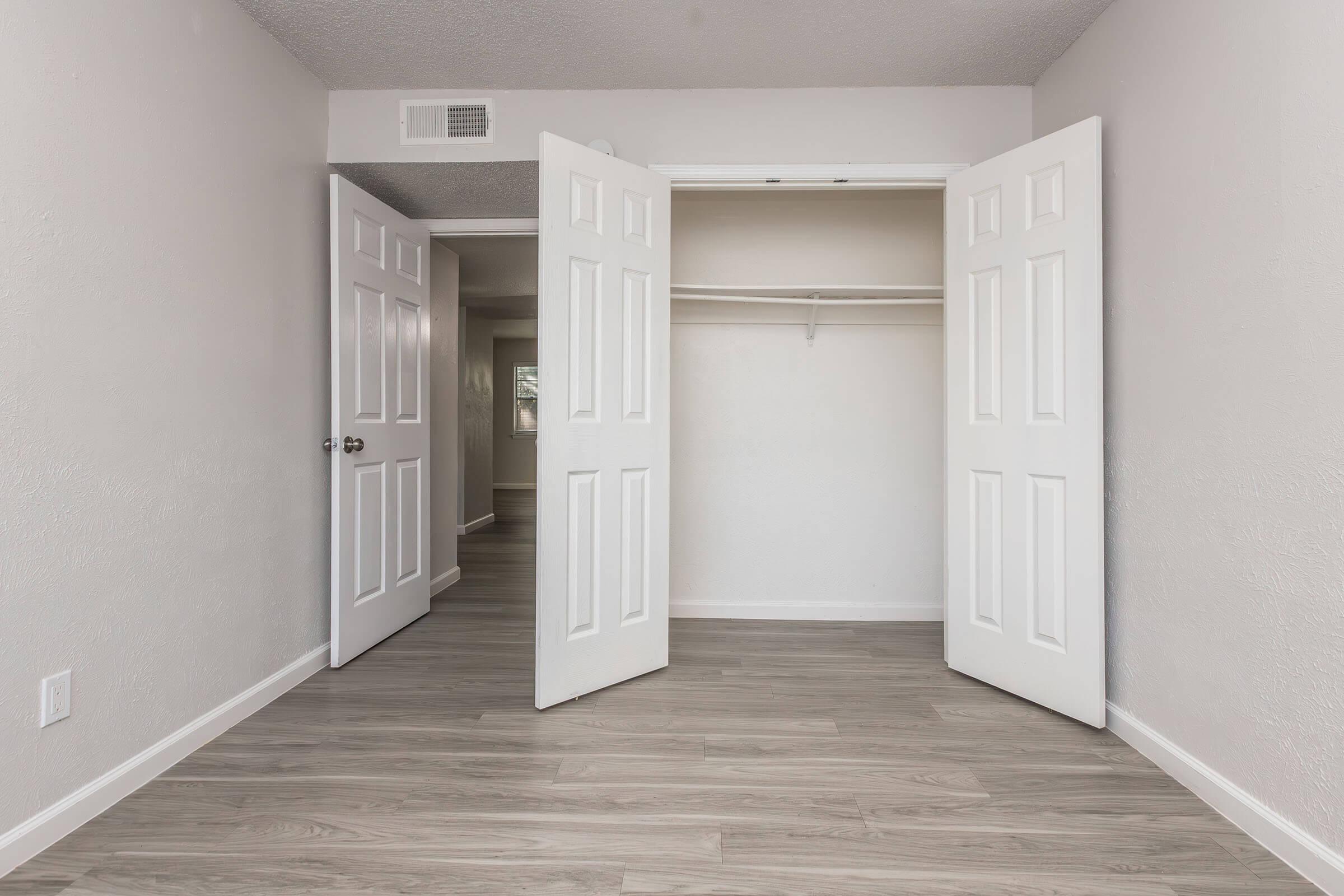 A spacious, empty room with light gray walls and a smooth, light wood floor. Two white double doors are open, revealing a closet with a single shelf. The room is well-lit, with another doorway visible in the background. The overall aesthetic is modern and minimalistic.