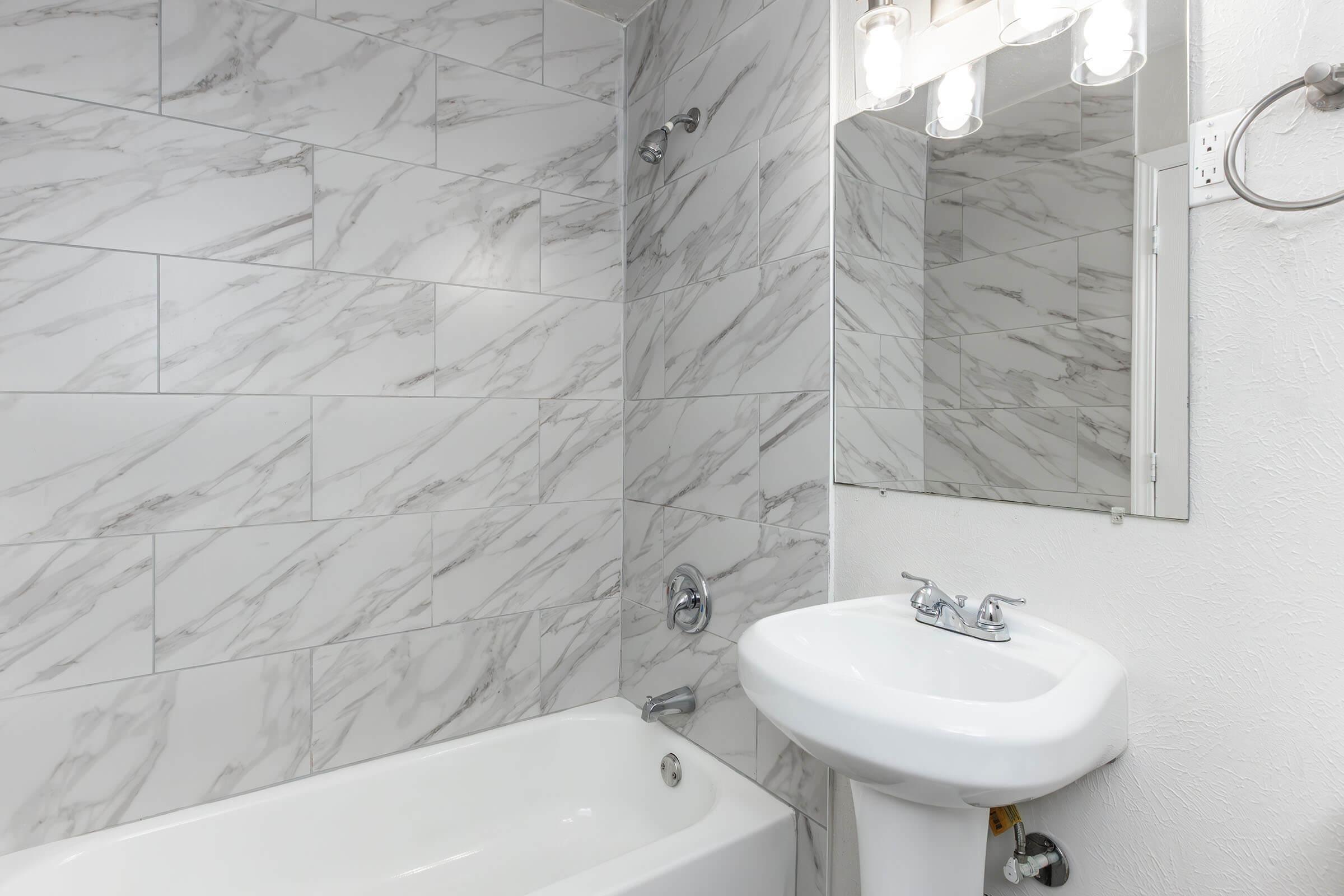 A clean, modern bathroom featuring a white tub, a marble-patterned tile wall, and a small sink with a faucet. Above the sink, there is a mirror and a light fixture with three bulbs. The overall design is sleek and contemporary, creating a bright and airy space.
