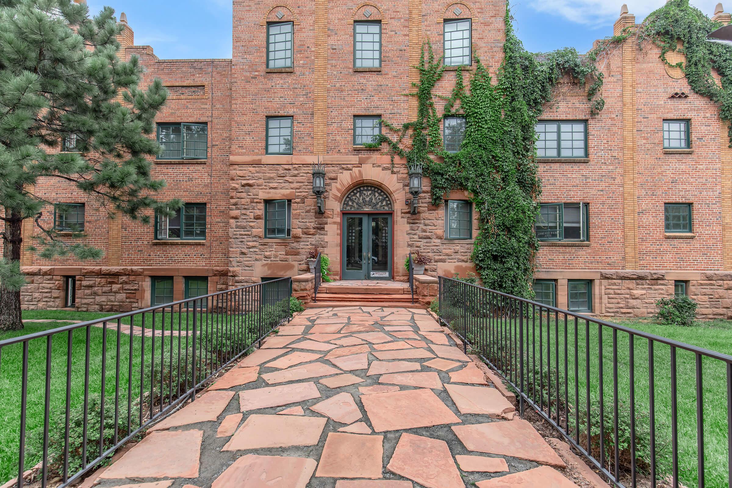A brick building with a decorative entrance, featuring large windows and ivy growing on the exterior. A stone pathway leads to the front door, flanked by manicured lawns and a few trees, creating an inviting entryway.