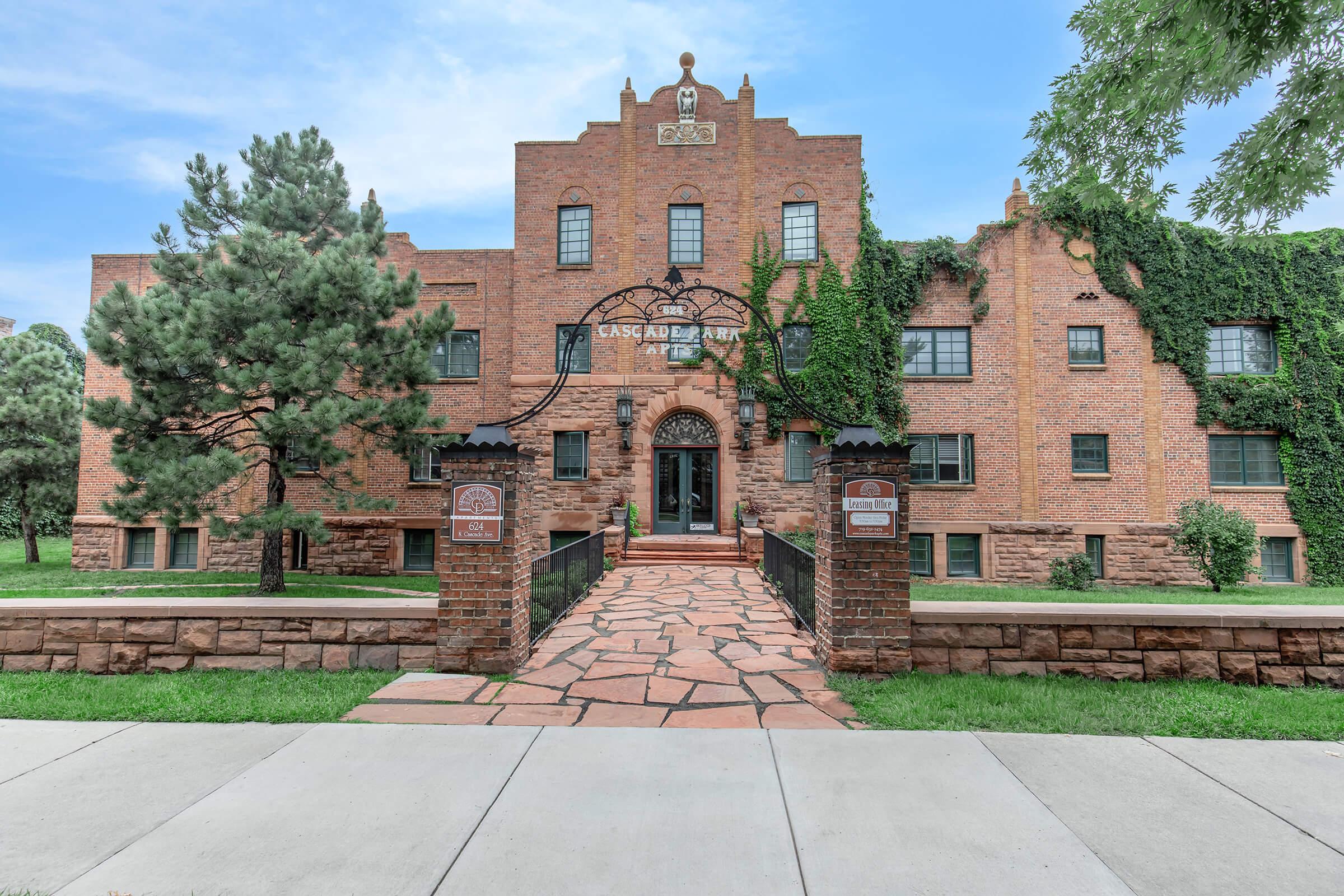 Historic brick building with ivy-covered walls, featuring arched entryways and a decorative facade. Mature trees are planted near the entrance, and a stone pathway leads to the front door. The sky is clear and blue, creating a serene atmosphere around the property.