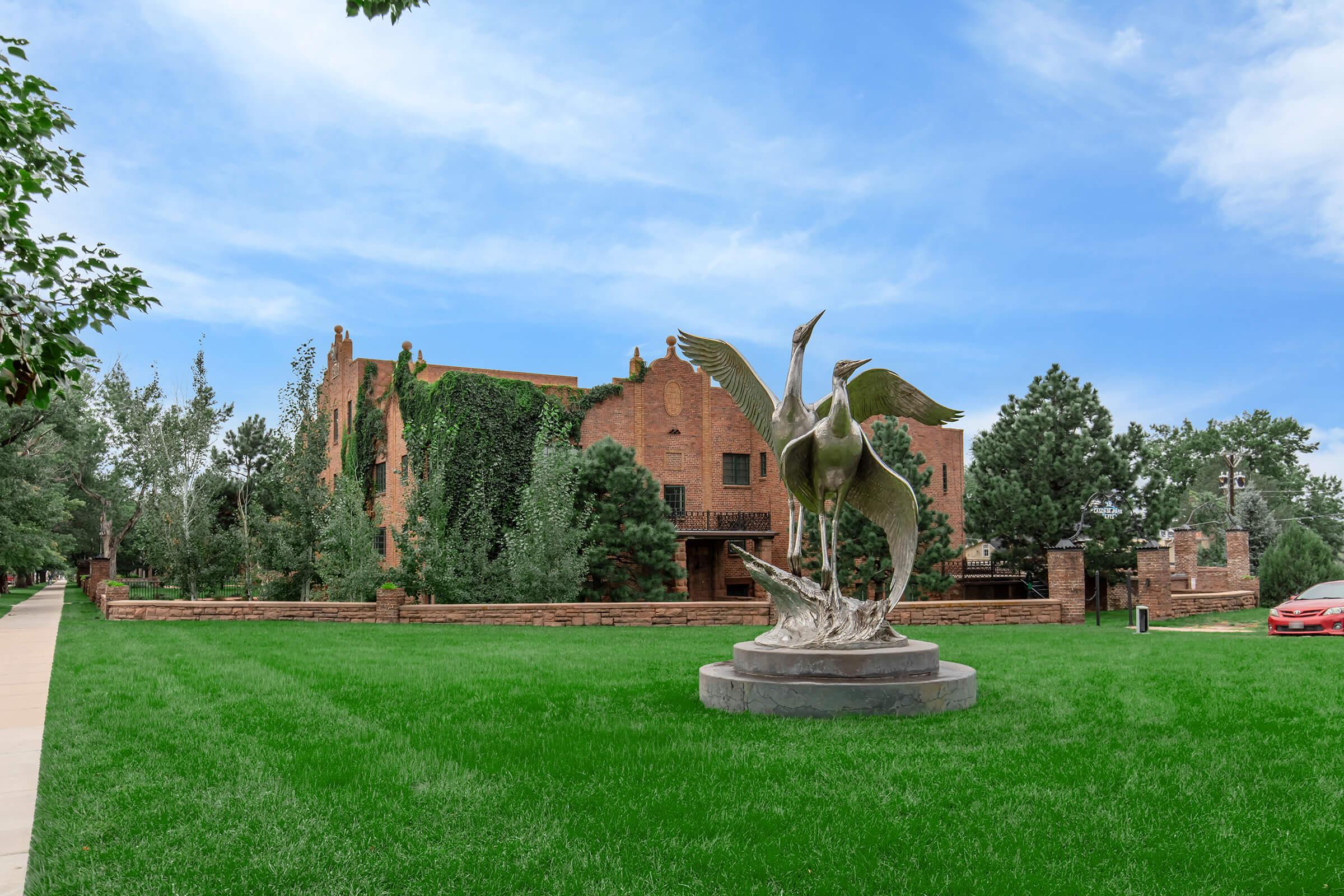 A bronze sculpture of two cranes stands on a circular base in a grassy area, with a brick building covered in ivy in the background. Trees are scattered around the lawn, and a red car is parked nearby under a blue sky with wispy clouds.