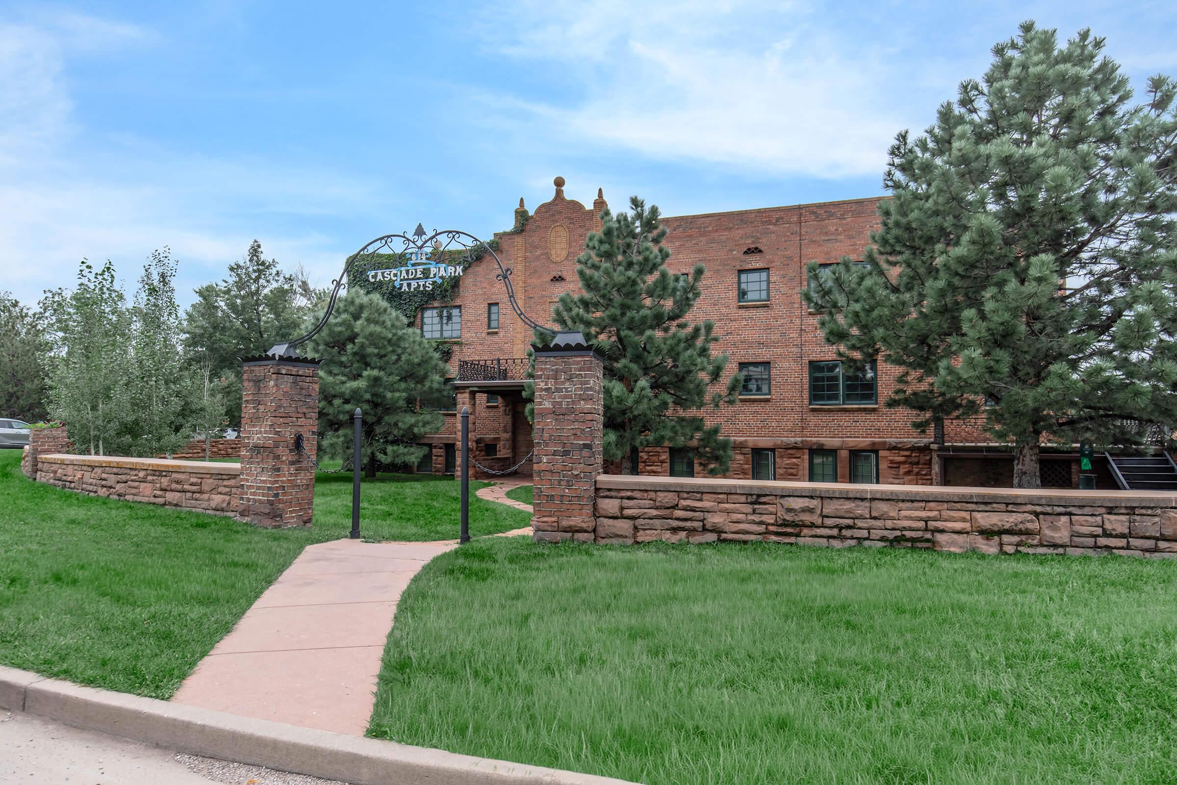 A red brick building with a decorative sign reading "Yesor Park" in front. The structure is surrounded by green grass and trees, with a pathway leading up to the entrance. The sky is partly cloudy, adding to the serene outdoor atmosphere.
