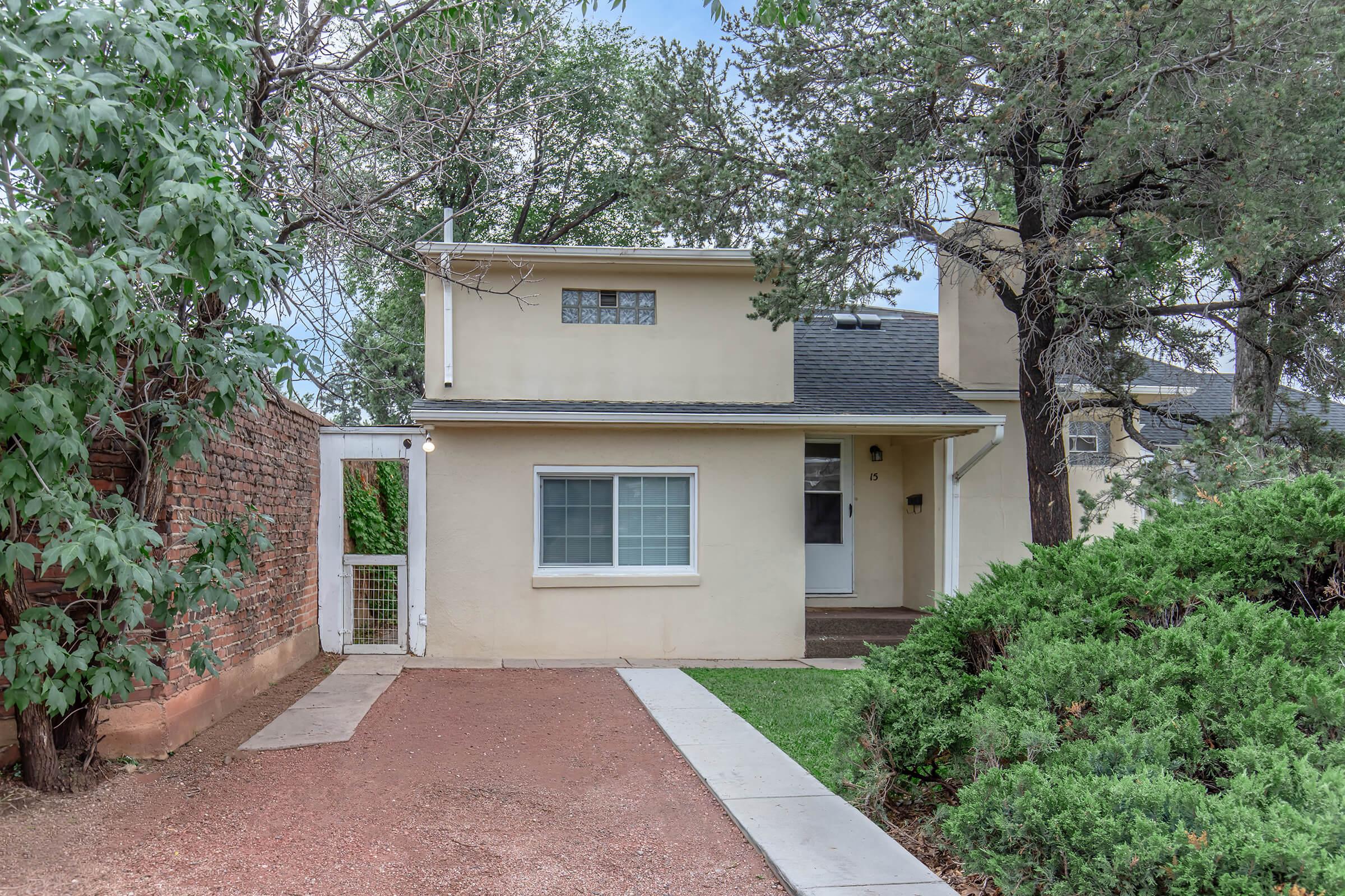 A two-story beige house with a small front entrance, surrounded by green trees and shrubs. A gravel path leads to the doorway. The house features a symmetrical design with a window above the entrance and a chimney on one side. A brick wall borders the left side of the property.