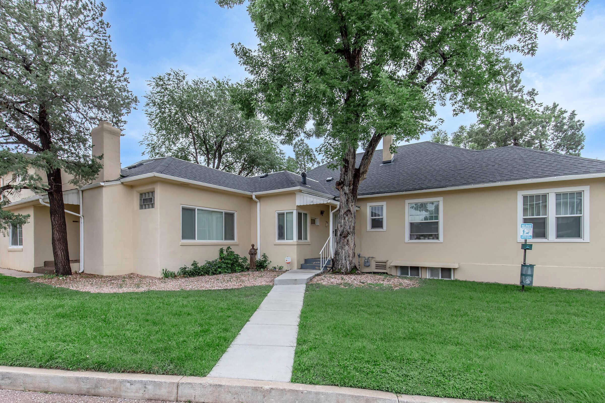A single-story home with beige exterior walls, a sloping gray roof, and multiple windows. The front yard features a manicured lawn bordered by gravel, with trees providing shade. A concrete walkway leads to the entrance, creating a welcoming path to the house.