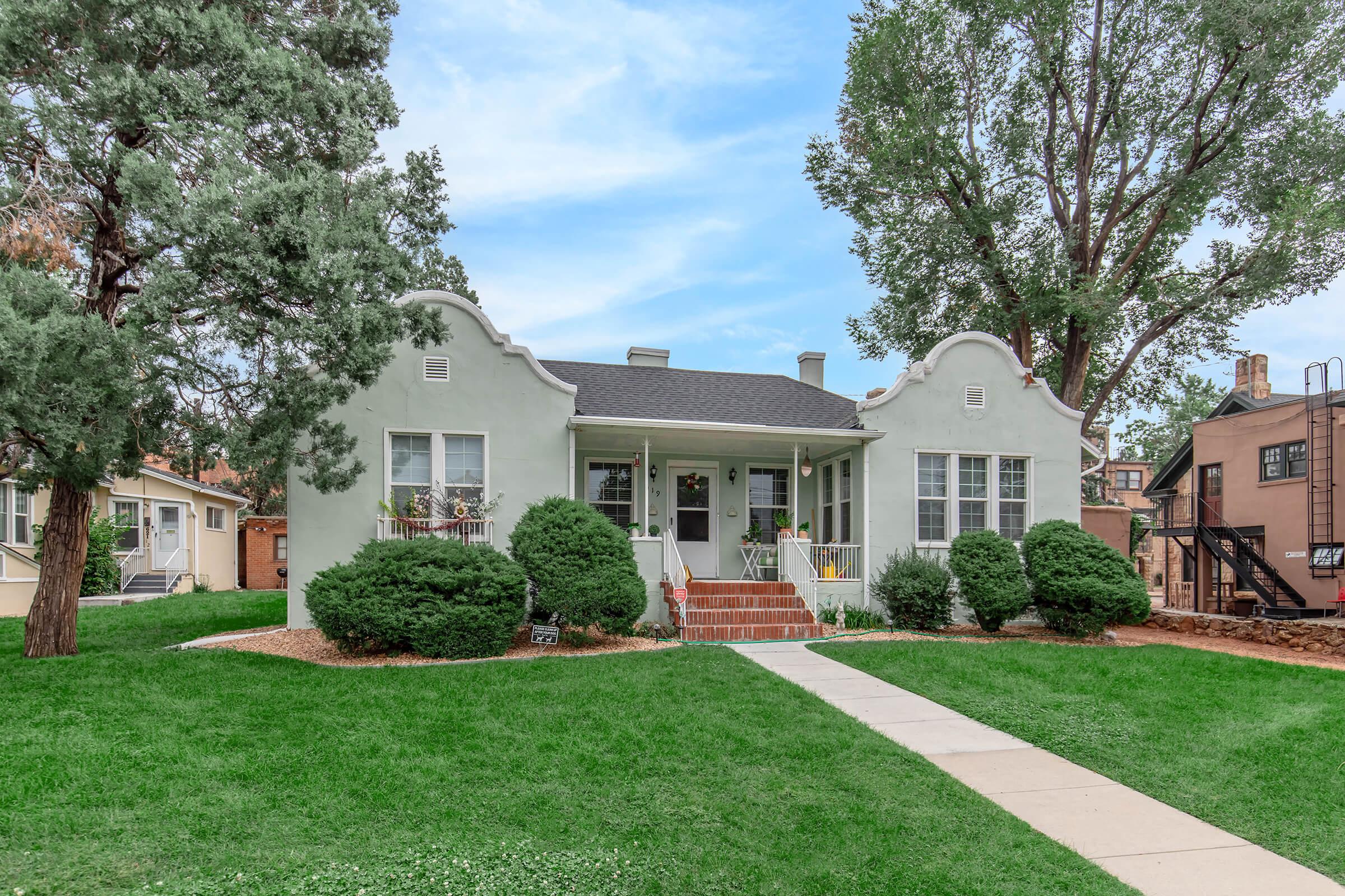 A charming single-story house with a pale green exterior, featuring multiple windows and decorative gables. There is a welcoming front porch with steps leading up to it. Lush green grass surrounds the house, complemented by neatly trimmed bushes and trees in the yard. A sidewalk leads up to the entrance.