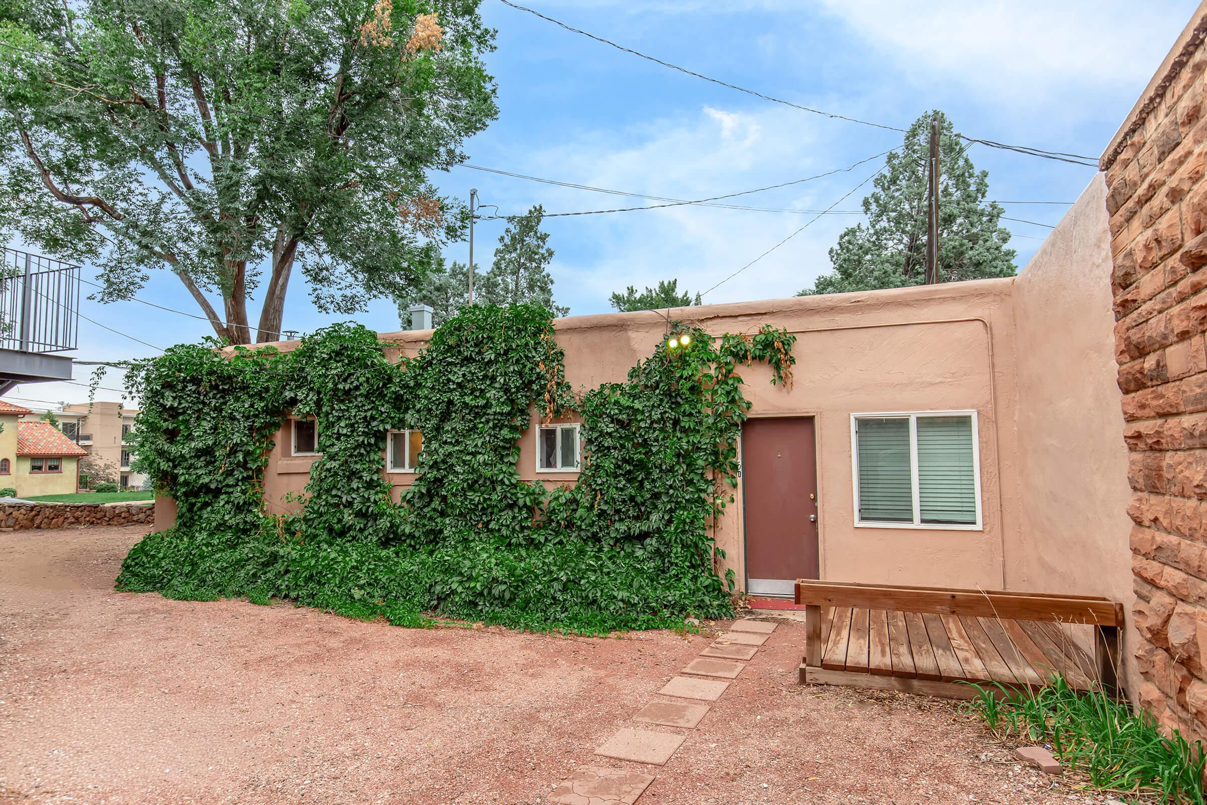 A single-story adobe-style building with a wooden door, partially covered in green vines. It features several small windows and is surrounded by gravel and a wooden bench. Trees and a blue sky are visible in the background, creating a serene and natural atmosphere.