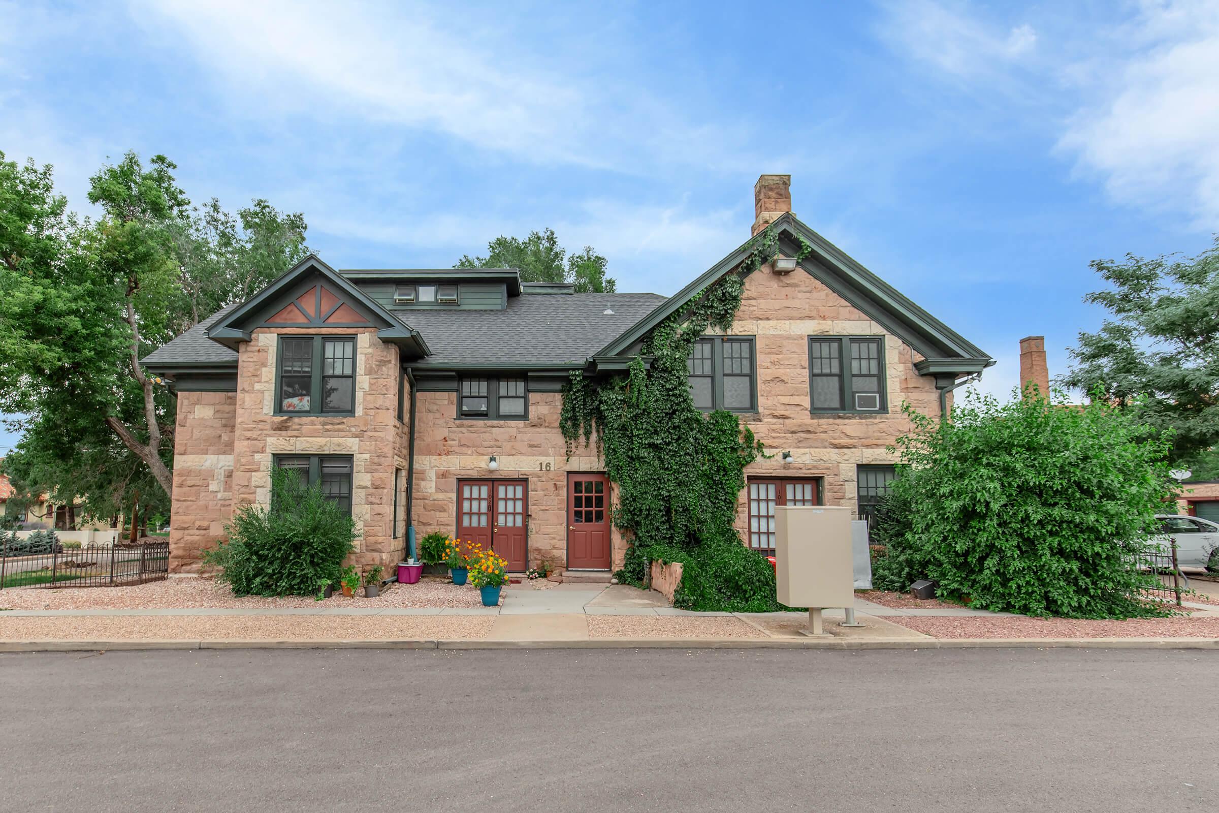 A charming two-story stone house with lush greenery and flowering plants around the entrance. It features a slate gray roof, large windows, and a welcoming front porch. The building is situated on a quiet street with a clear blue sky above, creating a picturesque neighborhood scene.