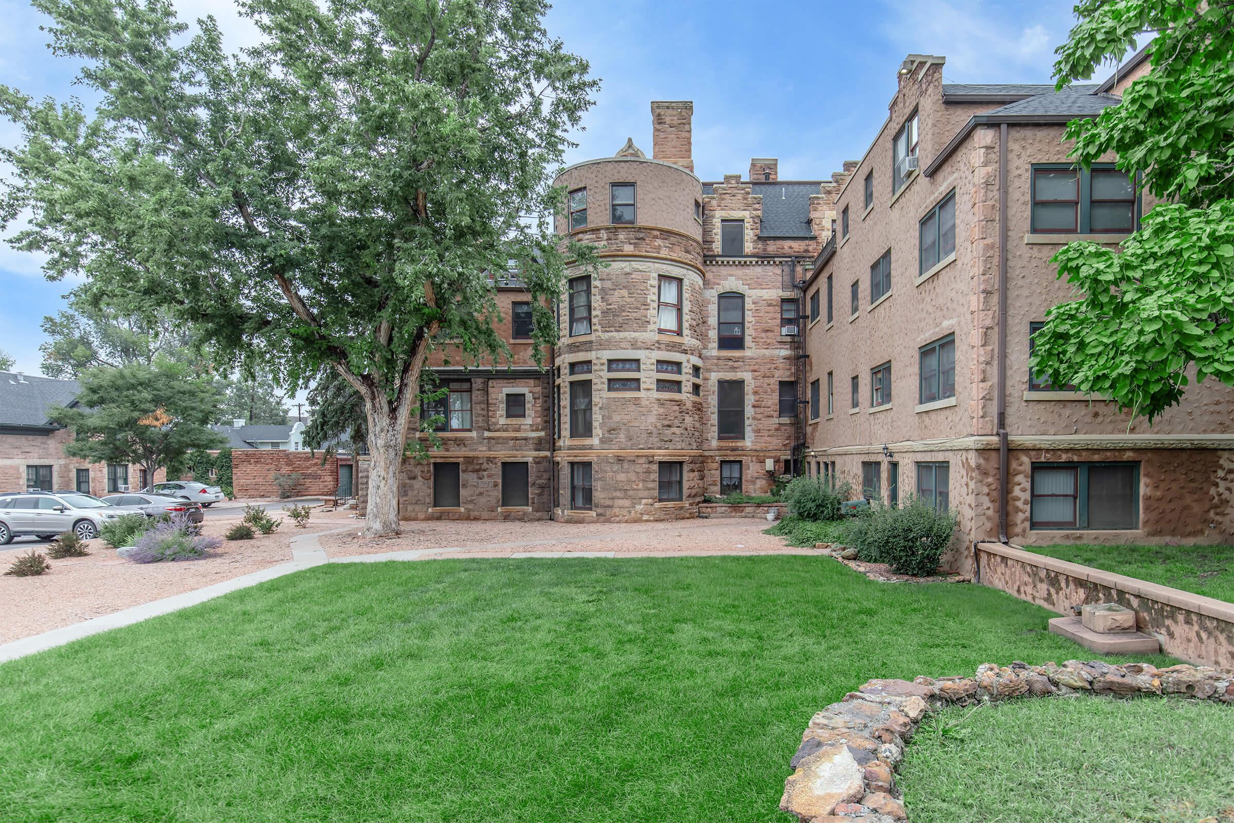 Historic building with a stone exterior, featuring a round turret and multiple windows. Surrounded by a well-maintained lawn, trees, and landscaped gardens. The architecture reflects a classic style, with visible details like brickwork and decorative elements. A peaceful, inviting atmosphere in a residential area.