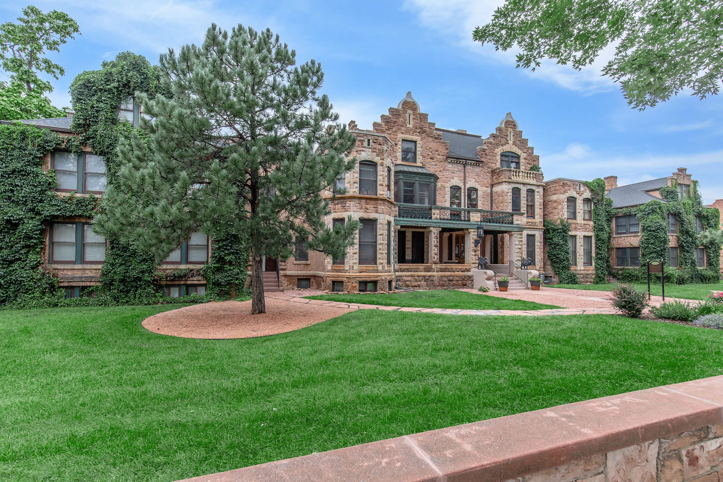 A large, historic stone mansion surrounded by lush greenery. The building features intricate architectural details, tall windows, and is partially covered in ivy. A well-kept lawn and landscaped pathways enhance the exterior, creating a welcoming atmosphere in a serene setting.