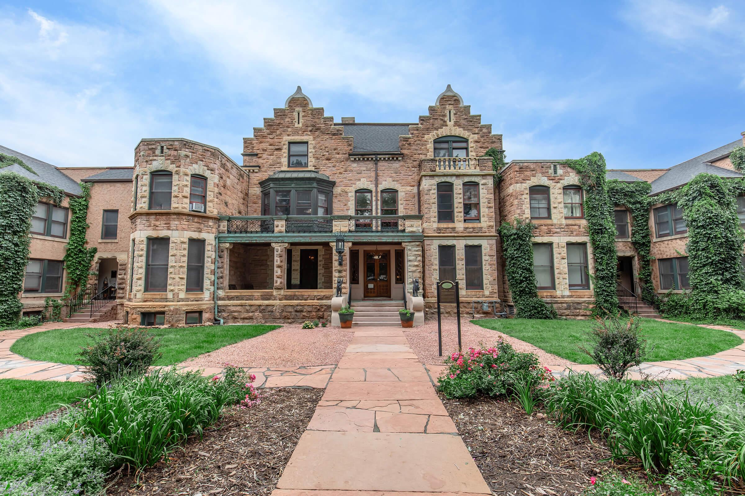 A grand stone mansion featuring intricate architectural details, surrounded by lush greenery and flowering plants. The front path is made of large stones, leading to the entrance, which has elegant windows and a balcony adorned with decorative elements. The sky above is bright and clear, enhancing the home's charm.