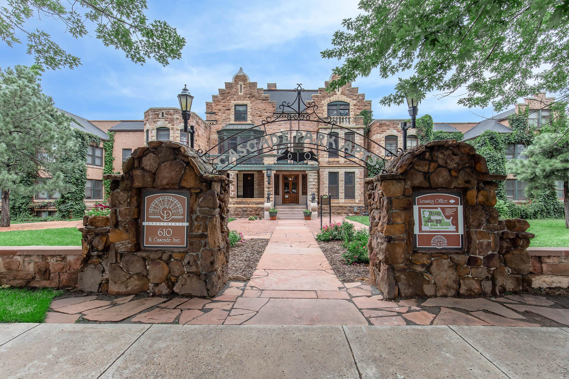 Entrance to a historic building with a stone archway and sign. There are landscaped green areas and trees flanking the entrance. A path made of stone leads to the main entrance of the building, which features a blend of architectural styles and inviting outdoor spaces.