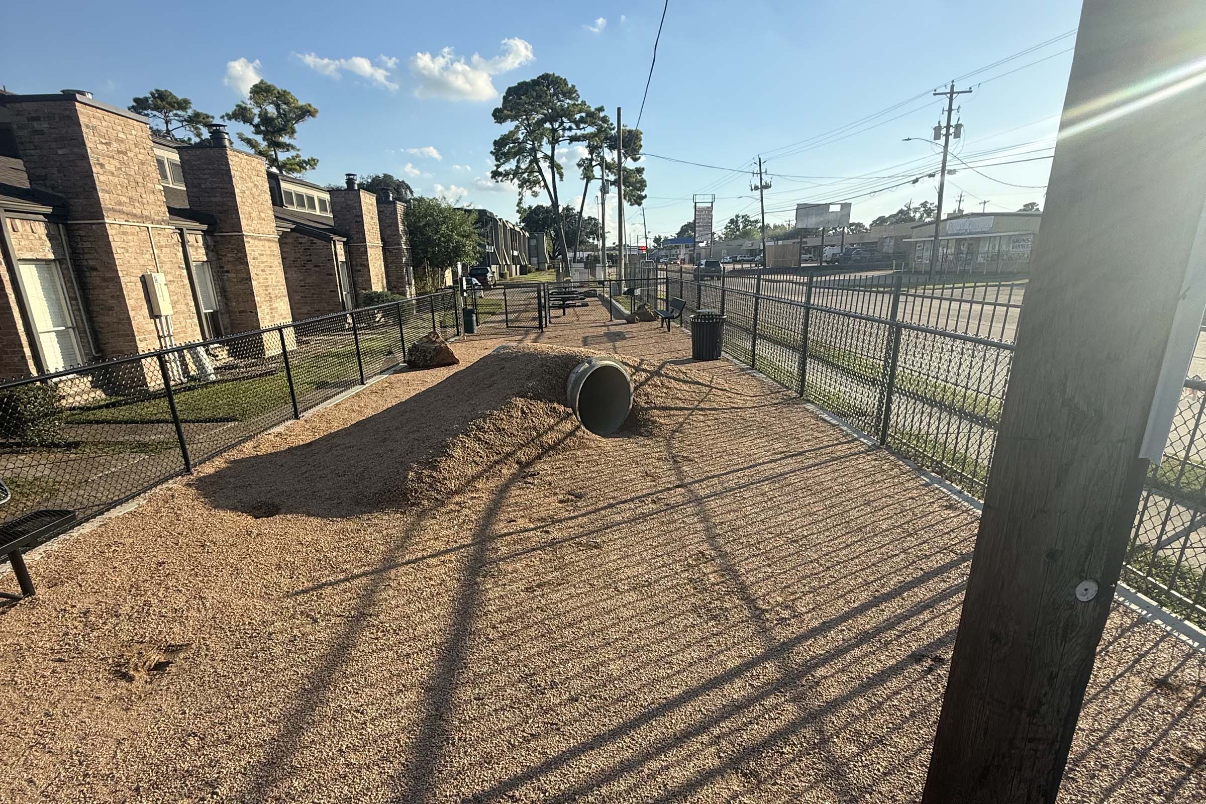 An empty dog park with gravel ground, featuring a large tube-like structure and a mound of dirt. Surrounding the area are fences, benches, and nearby trees, with a view of a street and buildings in the background. The scene is illuminated by afternoon sunlight.