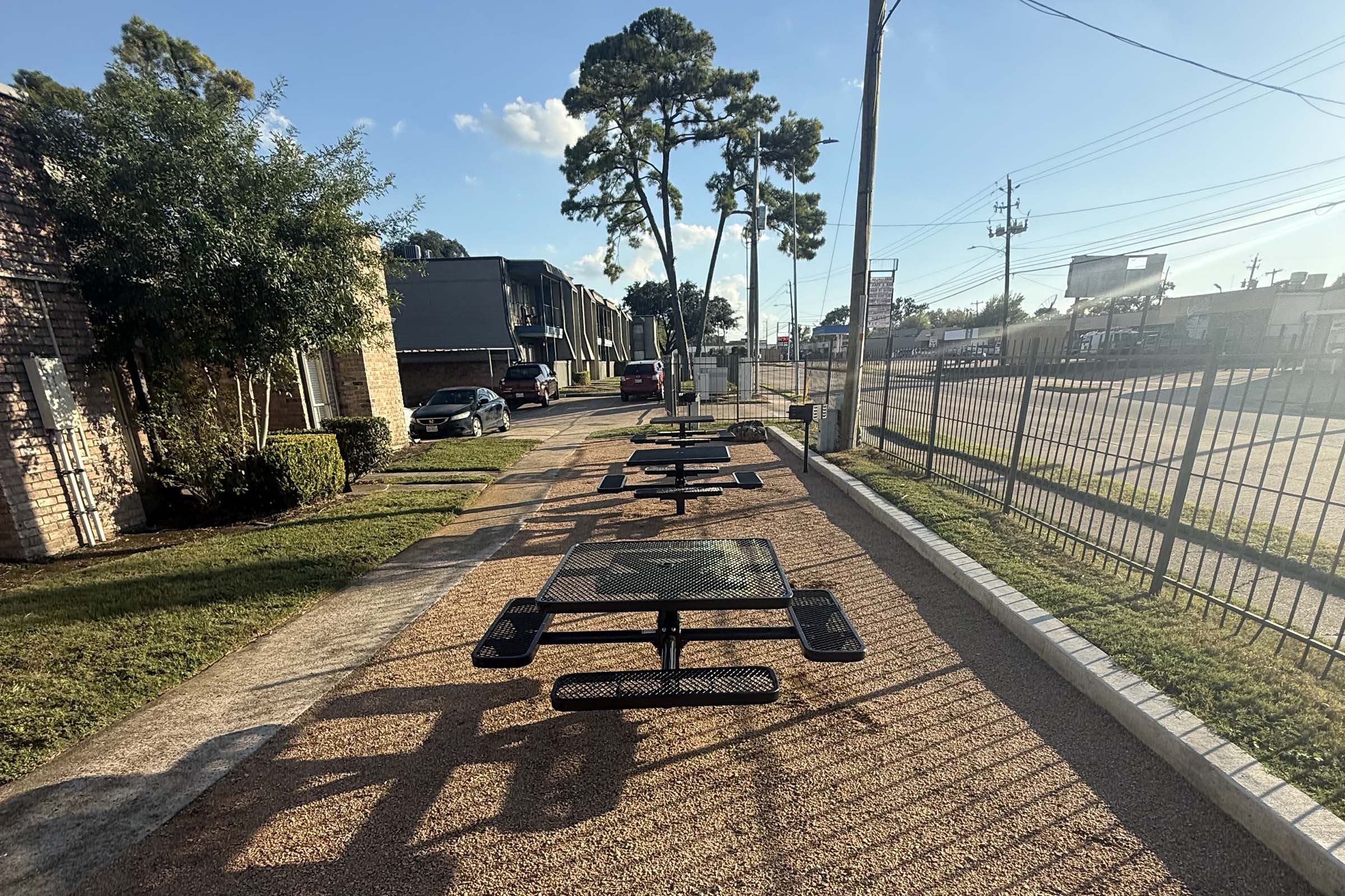 Rows of metal picnic tables on a pathway, surrounded by grass and shrubs. In the background, there are buildings and parked cars. The scene is well-lit, with a clear blue sky and a few clouds visible. A fence lines one side of the pathway, creating a separated outdoor space.