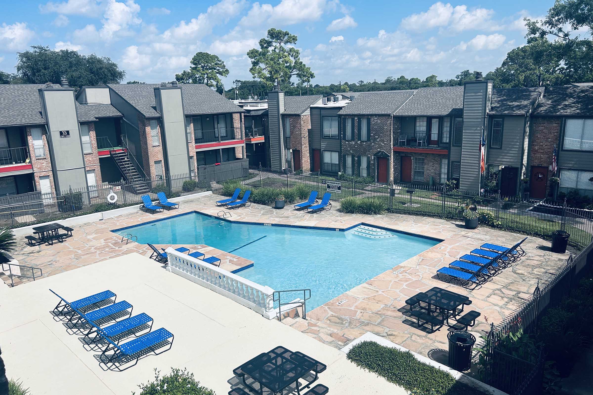 A bright outdoor scene featuring a rectangular swimming pool surrounded by lounge chairs and tables. The area is enclosed by low fencing, with two-story apartment buildings in the background, greenery, and a clear blue sky dotted with clouds.