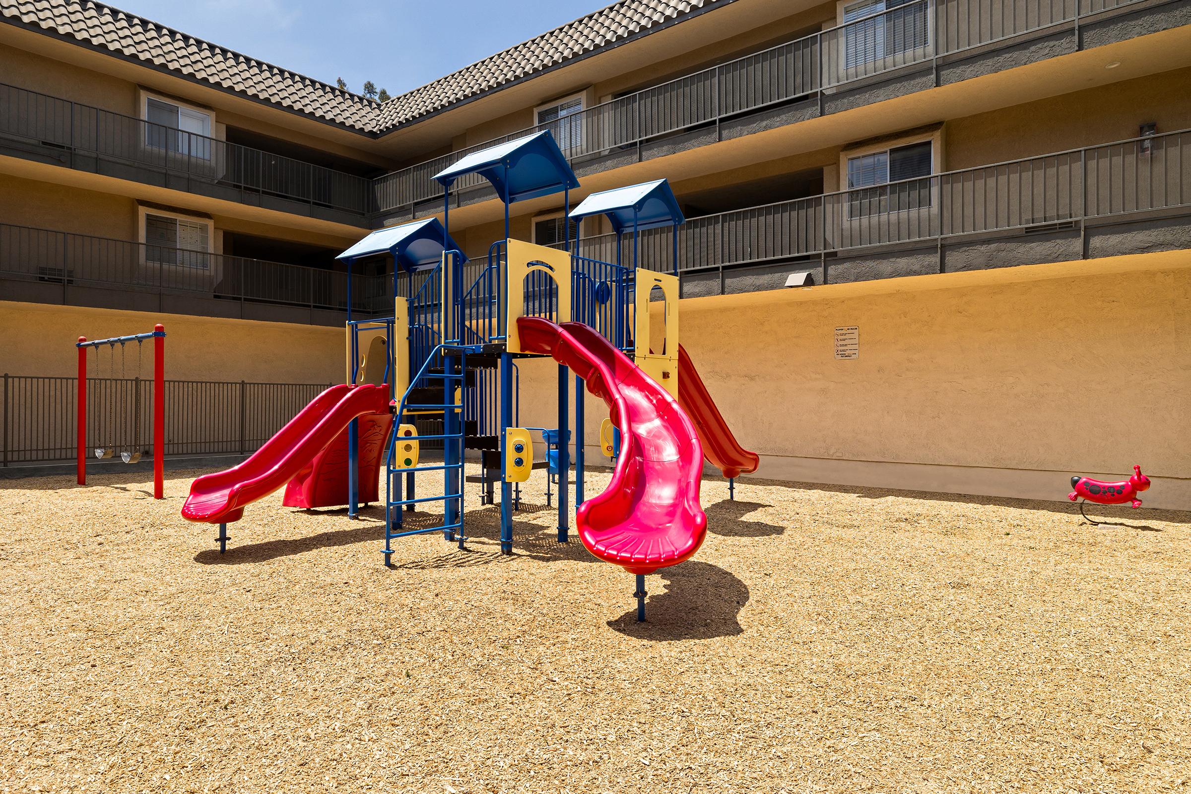 Colorful playground equipment featuring red slides, climbing structures, and a swing set, situated on a sandy surface within a courtyard surrounded by apartment buildings. The area is sunny and inviting, providing a space for children to play and enjoy outdoor activities.