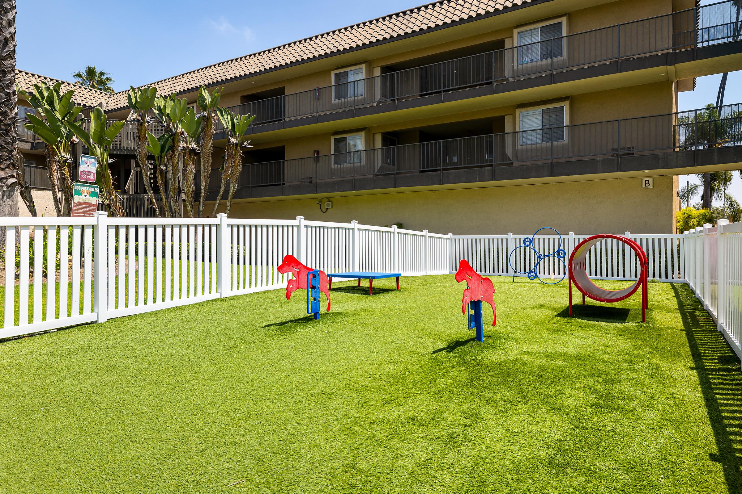 A small, fenced play area featuring artificial grass. Brightly colored play equipment includes two horse-shaped seesaws, a circular tunnel, and surrounding palm trees. In the background, a two-story building with balconies is visible under a clear blue sky.