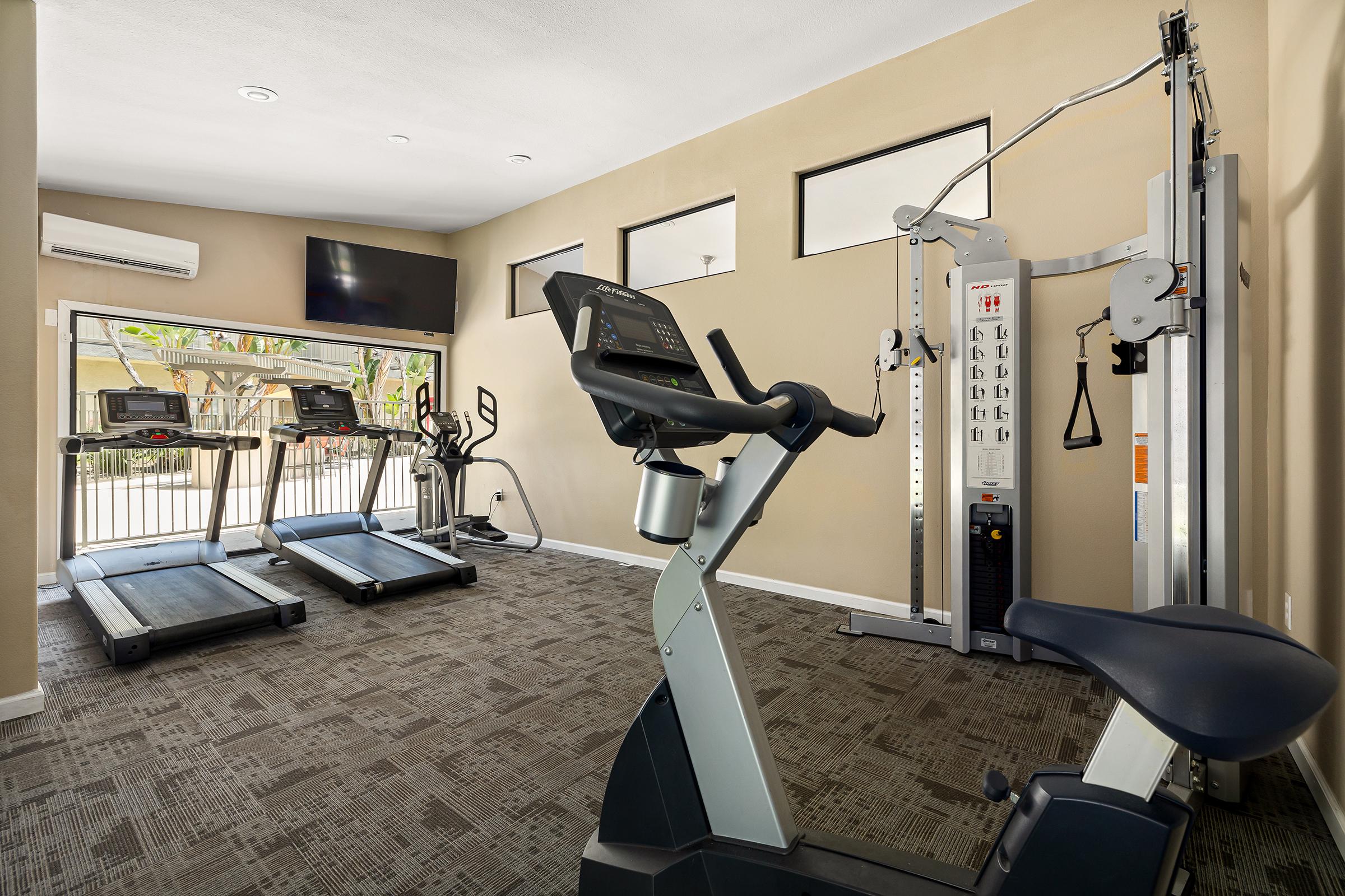 A modern fitness center featuring various exercise equipment. In the foreground, an upright bike is positioned, while two treadmills are visible in the background along with a multi-gym strength training machine. Large windows allow natural light into the space, and a television is mounted on the wall.