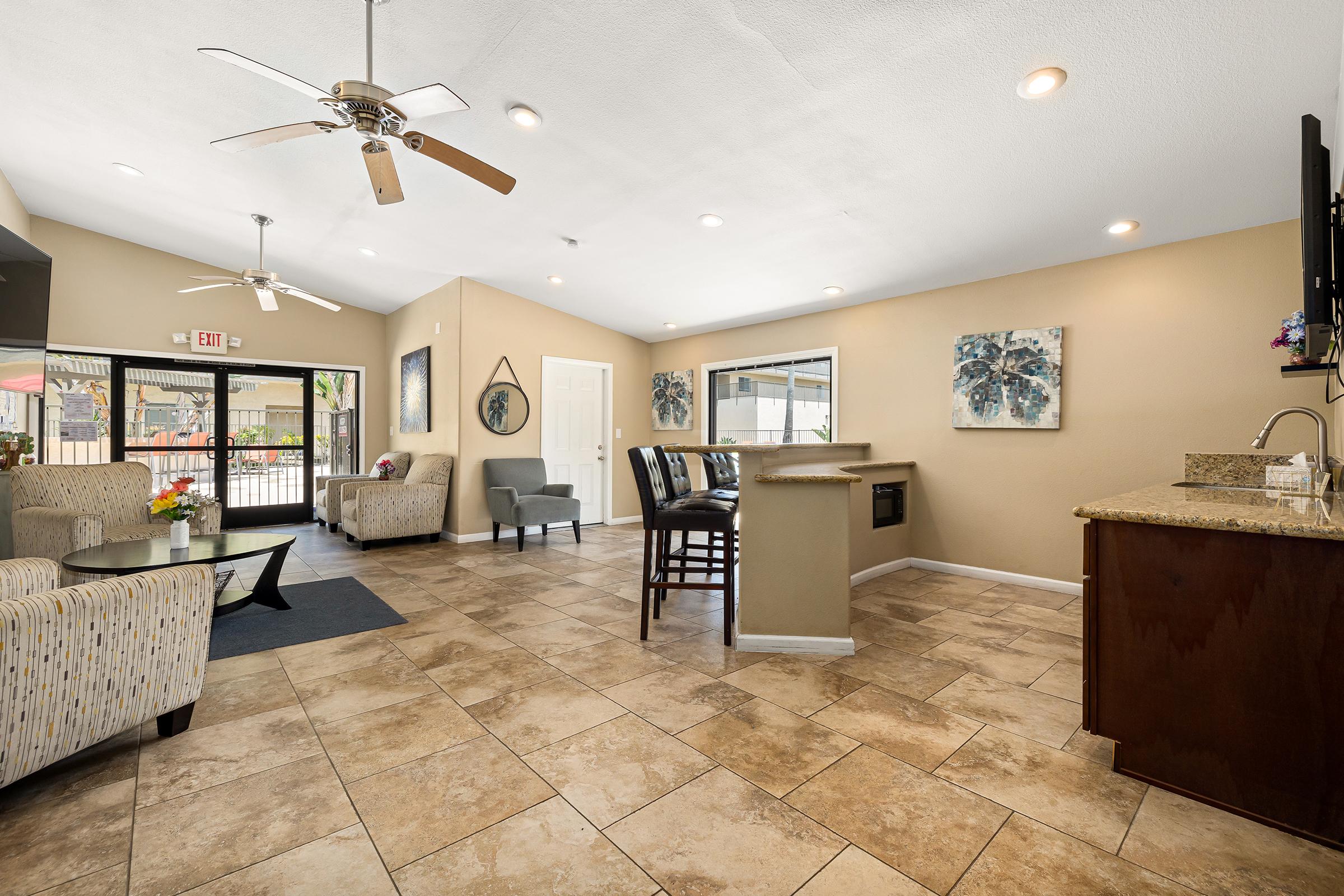 A spacious indoor living area featuring comfortable seating with colorful patterns, a granite countertop bar, and tile flooring. The space is well-lit with ceiling fans and large windows, showcasing a welcoming atmosphere with artwork on the walls.