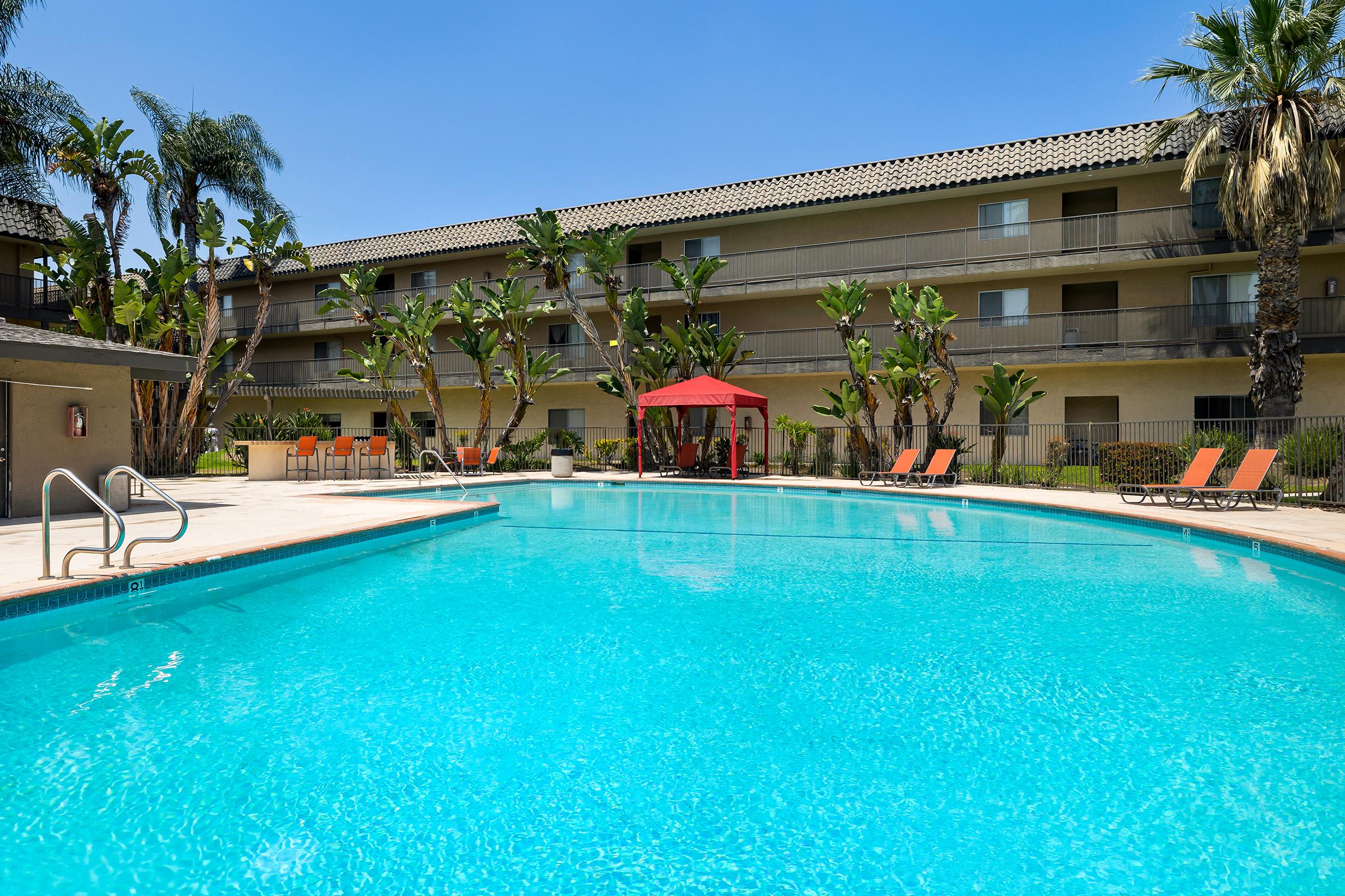 A clear blue swimming pool surrounded by lush palm trees and potted plants. In the foreground, there are orange lounge chairs and a red gazebo. The background features a three-story hotel with balconies, under a bright blue sky. The scene conveys a relaxing vacation atmosphere.