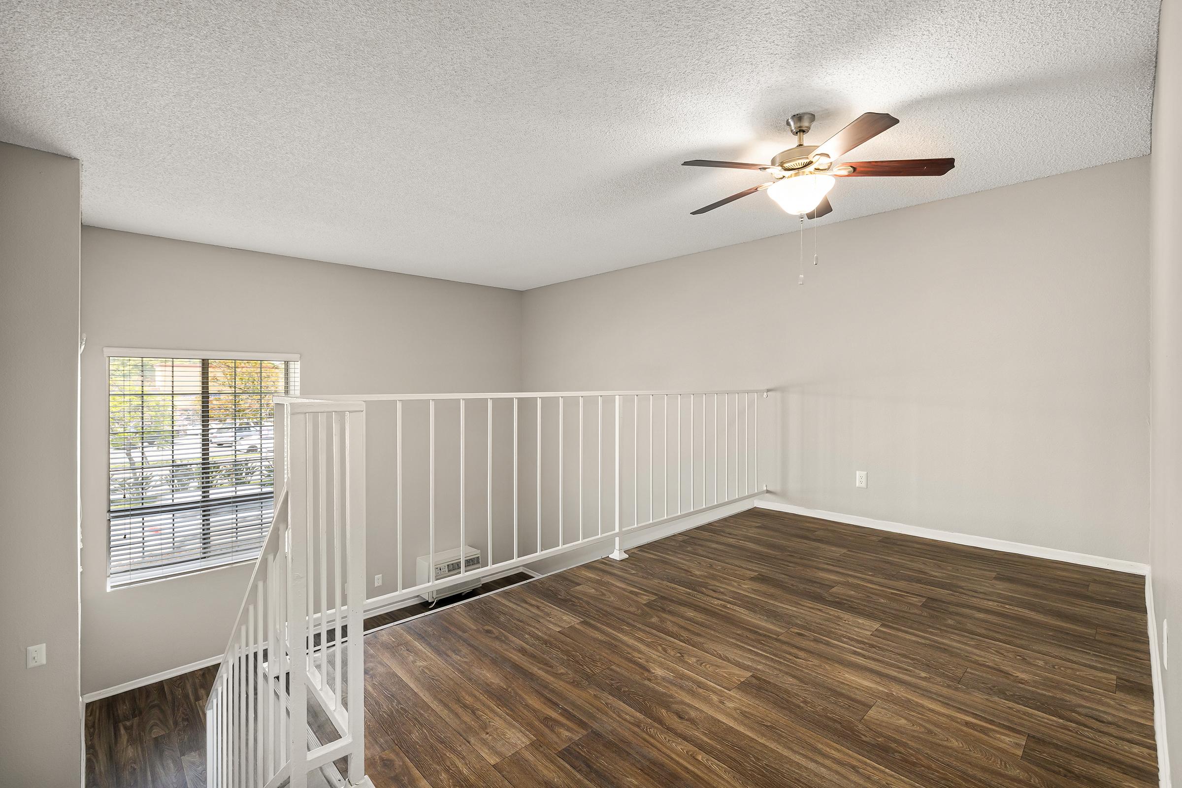 A well-lit, spacious room with a ceiling fan, featuring light-colored walls and wooden flooring. The room has a white railing partition and a window allowing natural light. It appears to be an upper level, open space, possibly designed for a living area or office.