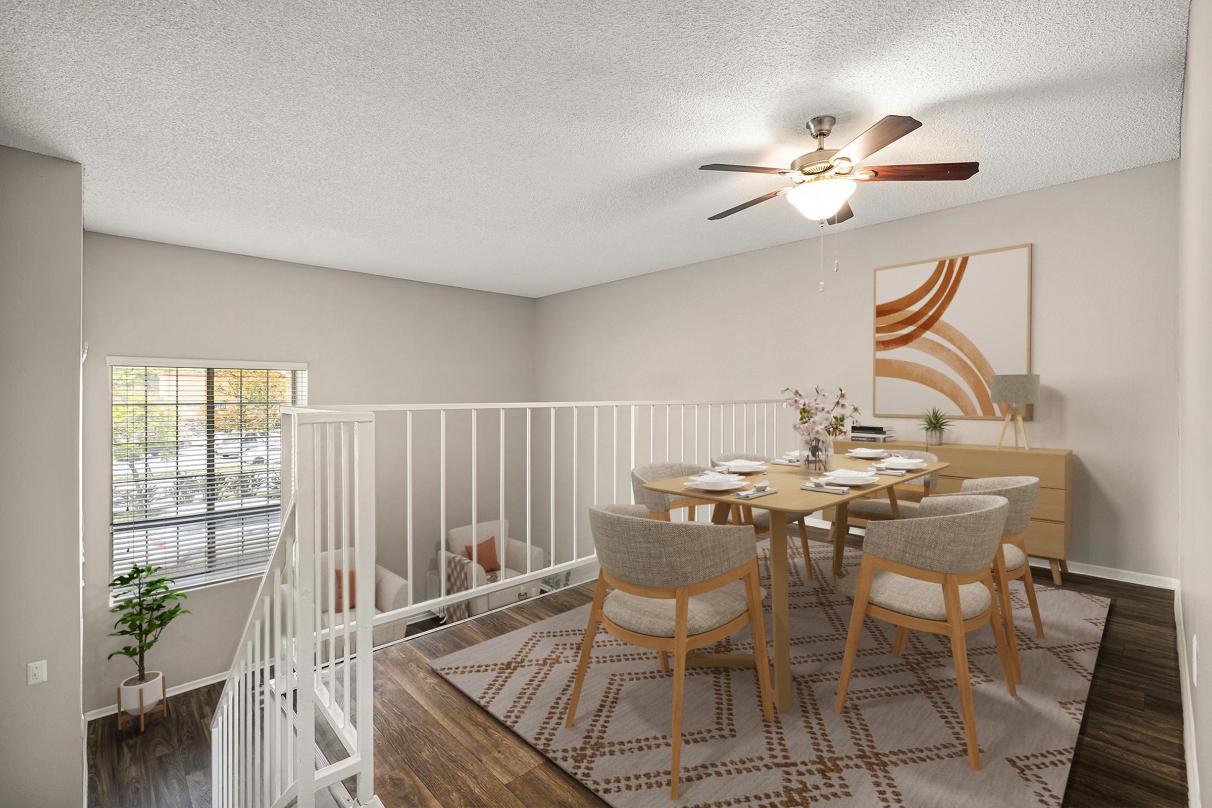A well-lit dining area featuring a neutral color palette. It includes a wooden table set for six, surrounded by upholstered chairs, a ceiling fan, and a decorative rug. A console table with decorative items is present, along with a large wall art piece. A window provides natural light, and a small plant adds a touch of greenery.