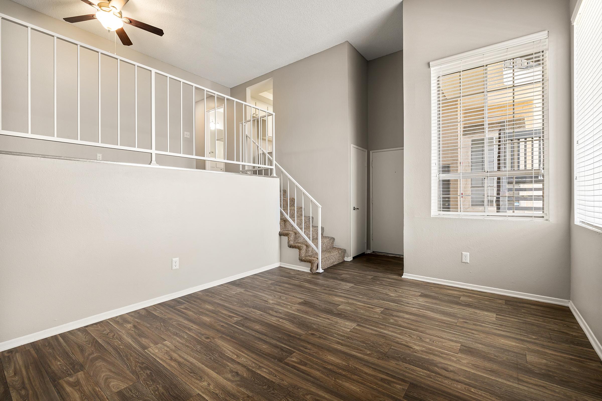 Interior of a modern, well-lit living space featuring light-colored walls, wooden flooring, and a staircase. A ceiling fan is visible, and there are large windows with blinds allowing natural light to enter. The space is open and airy, suitable for furniture arrangement and decoration.