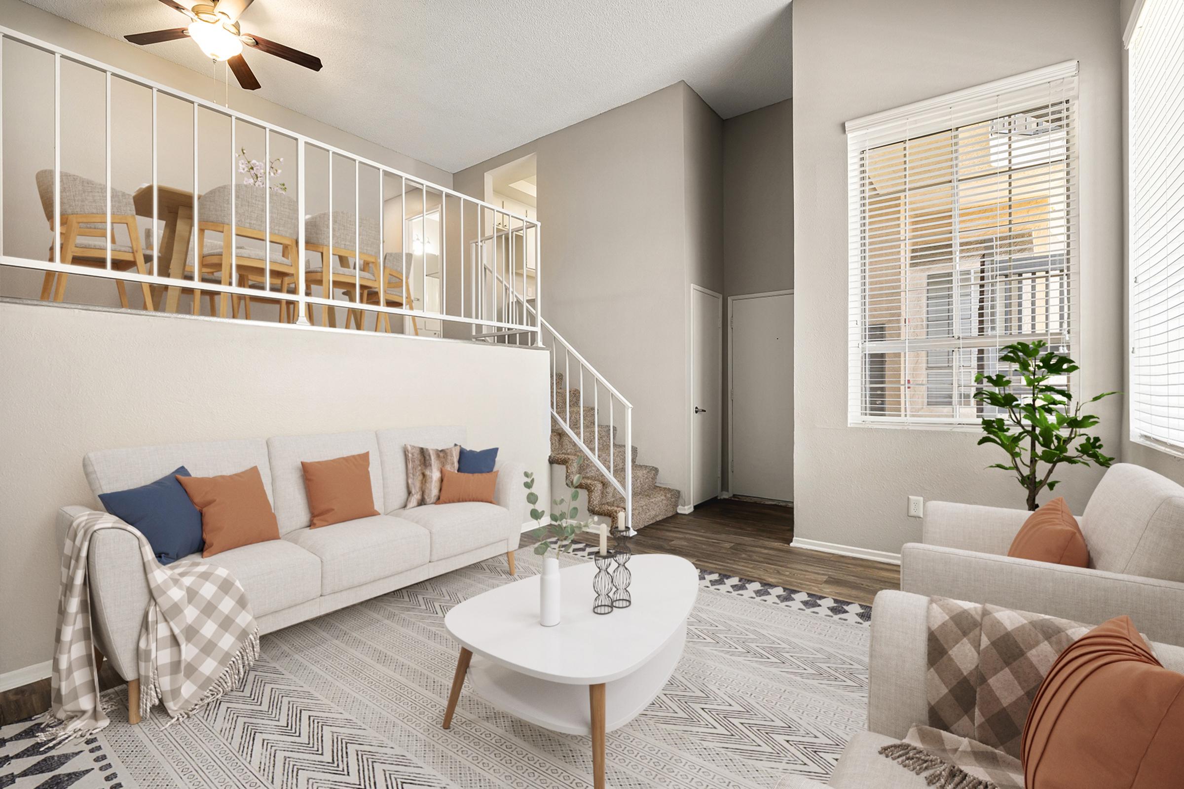 A modern living room featuring a light-colored sofa with orange and beige cushions, a stylish coffee table, and a cozy rug. A staircase leads to an upper level with dining chairs visible, while bright sunlight filters through large windows, creating a warm and inviting atmosphere.
