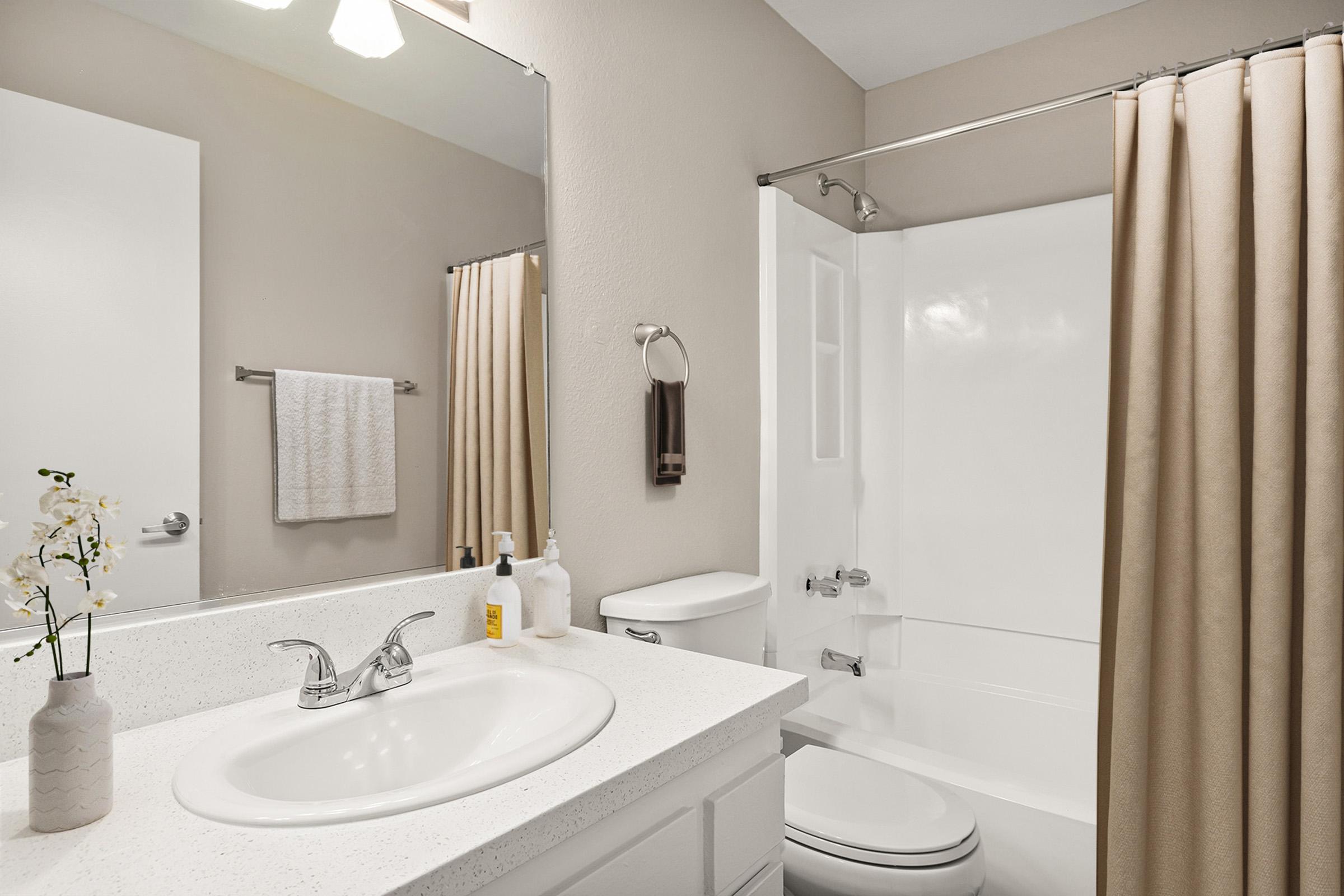 A clean bathroom featuring a white sink with a mirror above, a shower area with a beige curtain, a toilet, and a small vase with flowers on the countertop. Soft lighting enhances the neutral tones of the walls and fixtures.