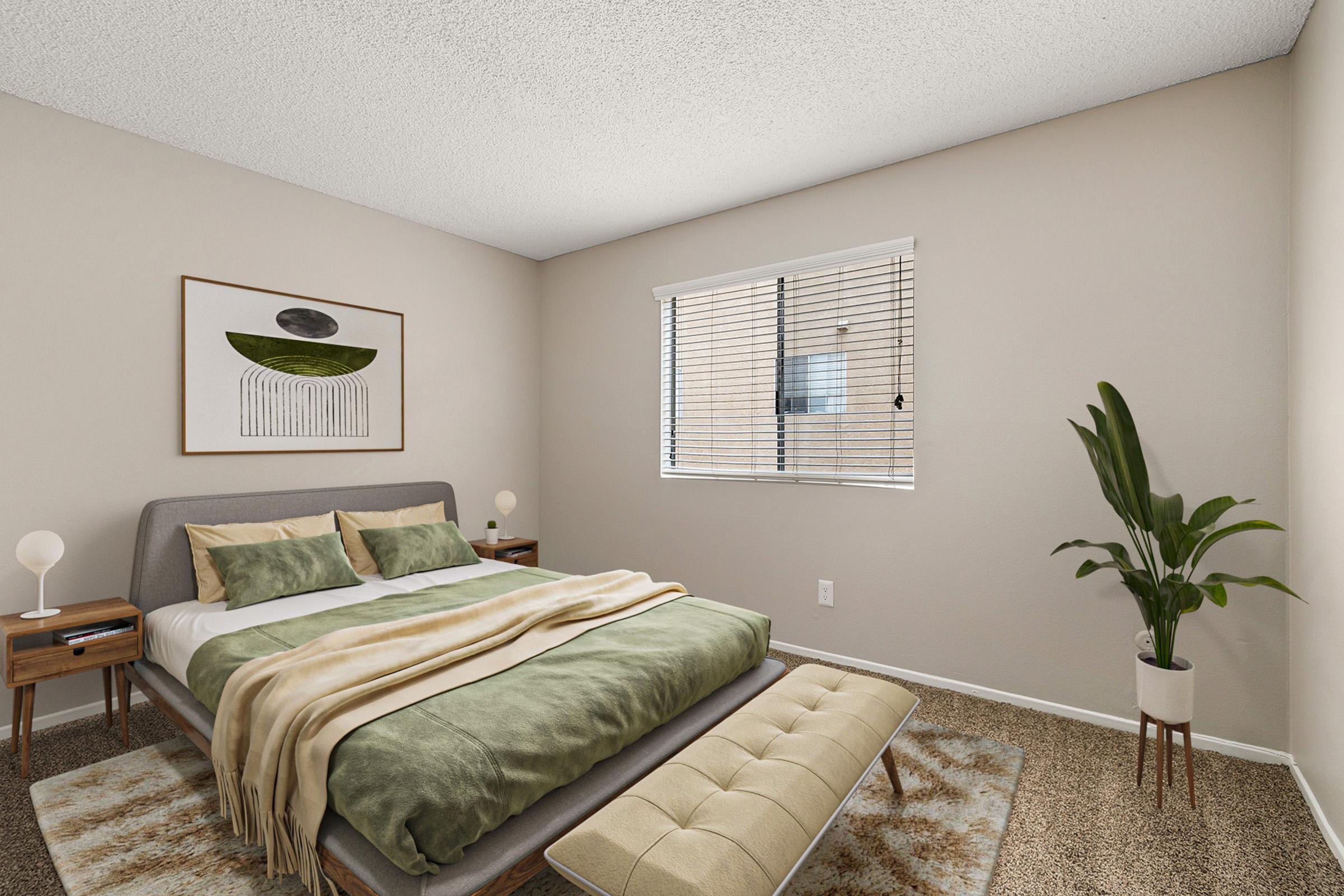 A cozy bedroom featuring a gray bed with green and white bedding, complemented by a tan throw blanket. Two bedside tables with lamps are on either side, and a large plant sits near the window. The room has light-colored walls and a textured carpet, creating a warm and inviting atmosphere.