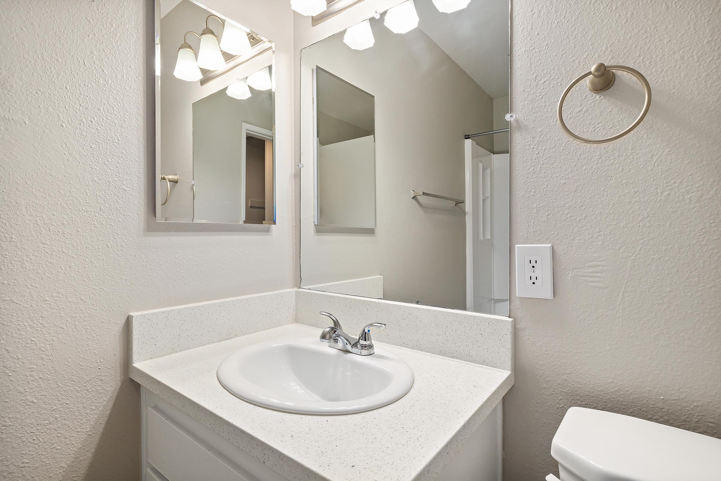 A clean and modern bathroom sink area featuring a white countertop, a single basin sink with dual chrome faucets, and two mirrors above. The walls are neutral in color, and there is a light fixture with three bulbs. A towel ring is mounted on the wall, and a toilet is positioned beside the sink.