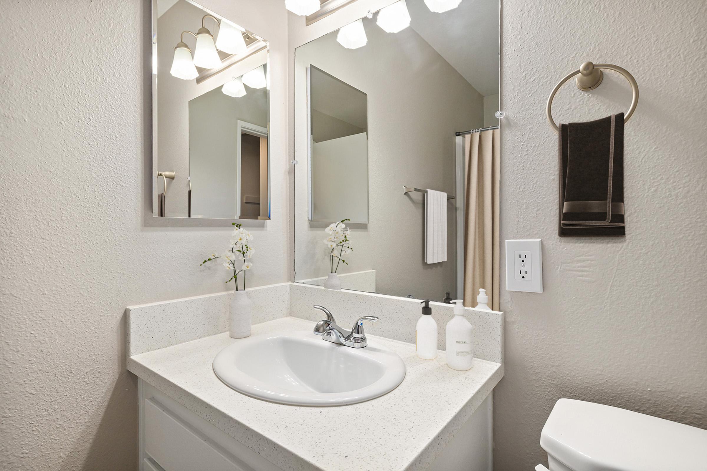 A clean, modern bathroom featuring a white sink with a polished countertop, a mirror above, and decorative flowers. Soft lighting from three overhead fixtures enhances the space. A towel holder with a dark towel and a soap dispenser are also visible, contributing to the tidy and inviting atmosphere.