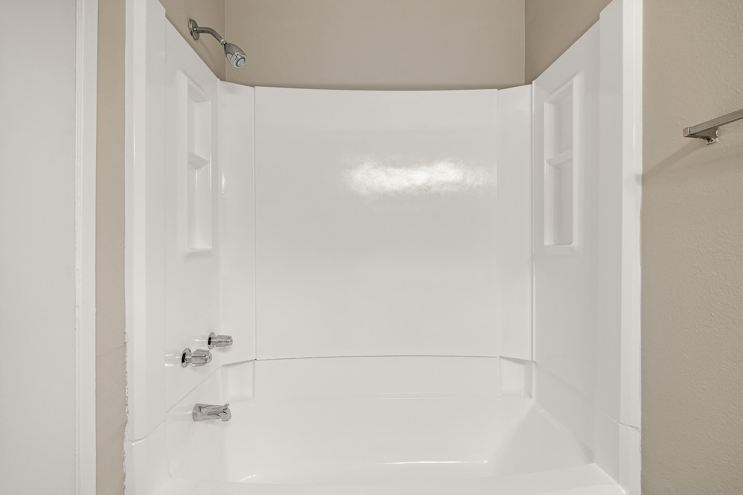 A clean, empty shower stall with white walls and a smooth, shiny floor. The space features a showerhead mounted on the wall and two faucet controls visible on the side. The overall design is modern and minimalist, with a light-colored wall surrounding the enclosure.