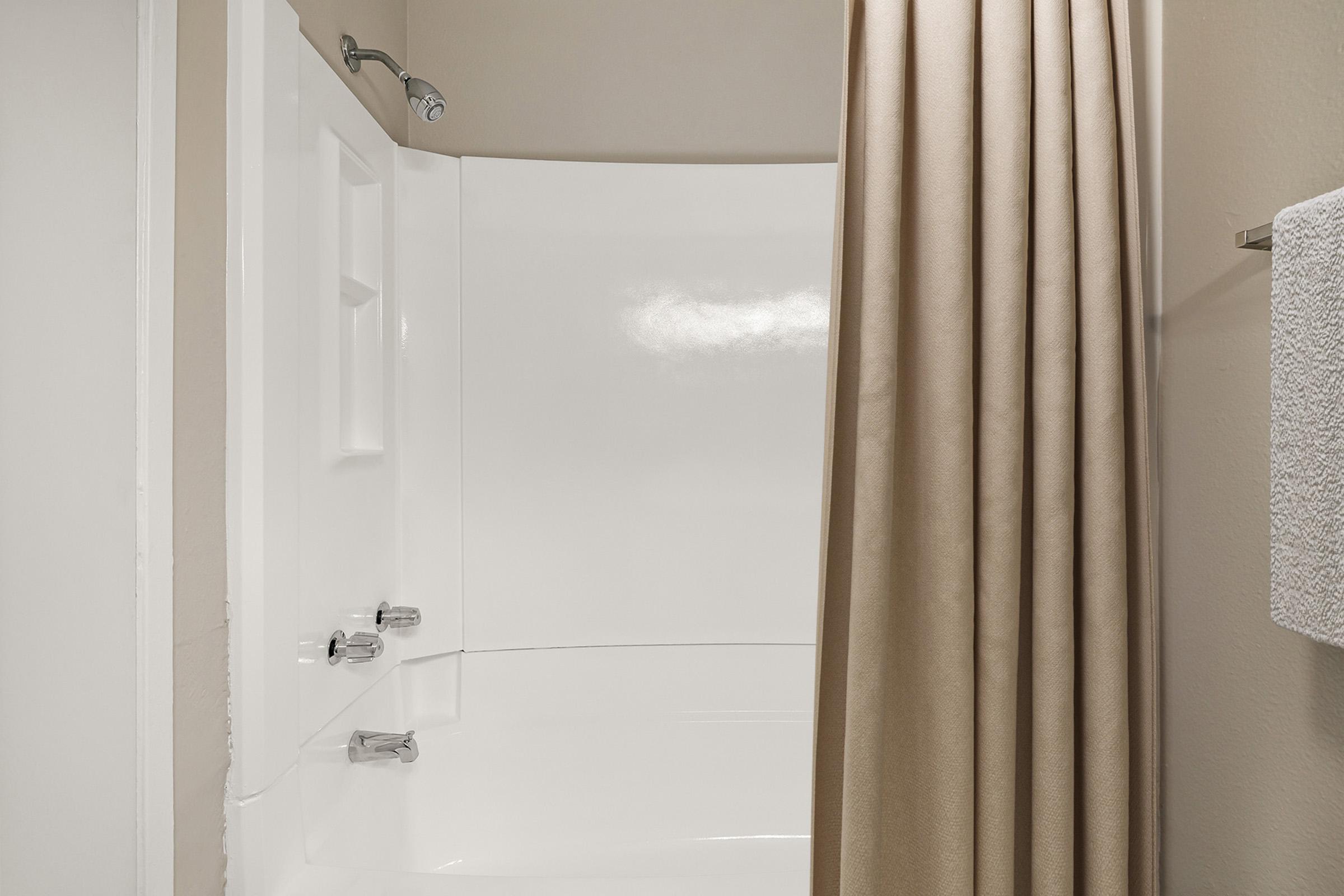 A clean and simple bathroom shower area. The shower features a white tub and curved shower wall, with a beige curtain hanging from a rod. A white towel is neatly hung on a towel rack nearby. The walls are painted in a neutral color, creating a calm and spacious appearance.